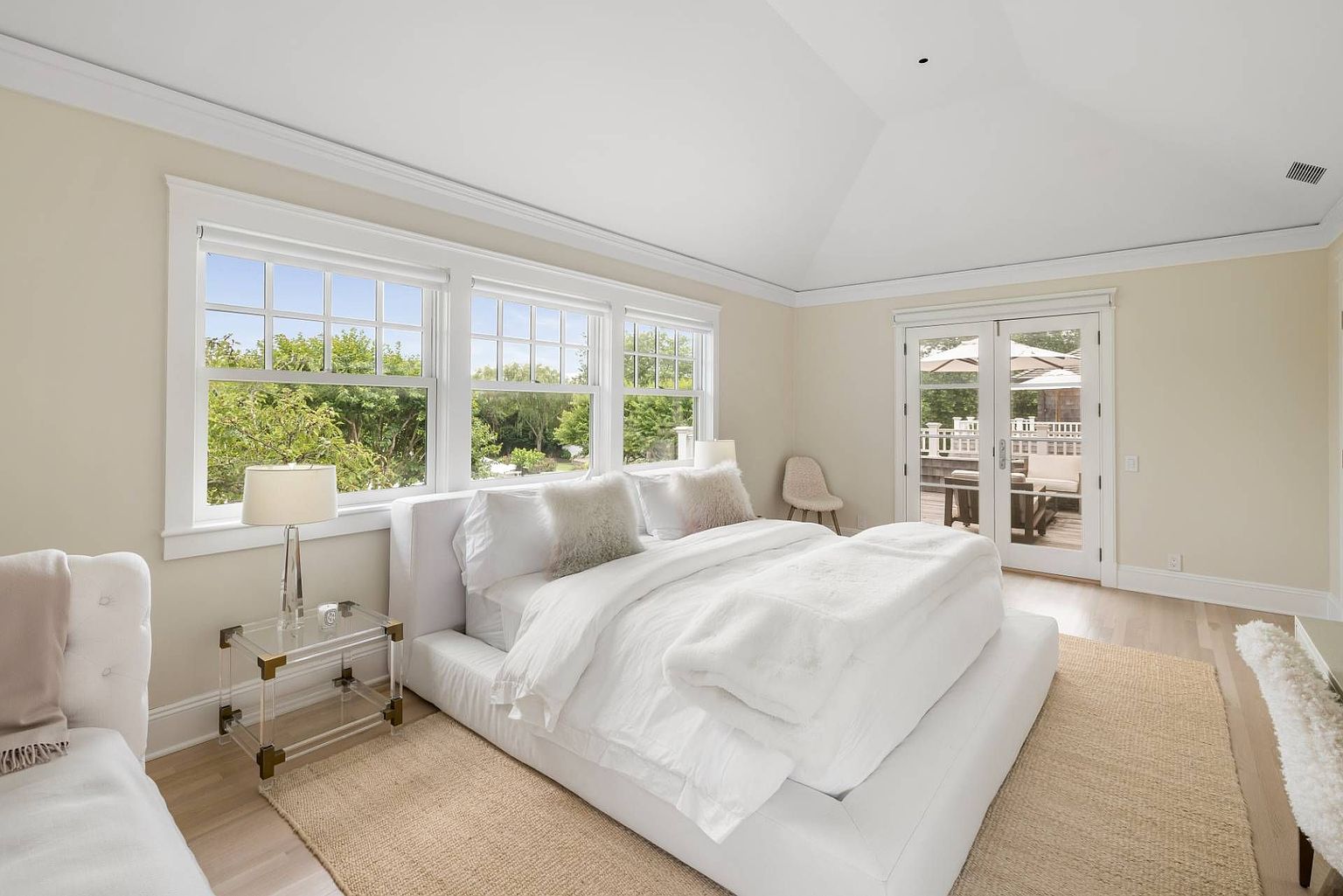 This bright and airy primary bedroom features a large, low-profile white upholstered bed centered in the room on a textured neutral rug. The space is illuminated by natural light streaming through a series of large windows and a set of glass-paned French doors that open onto an outdoor deck. The room is finished with light-toned wood flooring and a vaulted ceiling, creating a serene and spacious atmosphere.