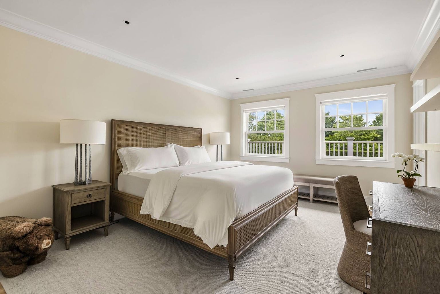 This primary bedroom features a large bed with a woven cane headboard, centered between two matching nightstands topped with modern table lamps. To the right, a desk area with a chair and a small potted plant provides a functional workspace, while a bench sits beneath two windows that offer a view of the outdoors. The room is finished with neutral-toned walls and light-colored carpeting, creating a serene and inviting atmosphere.