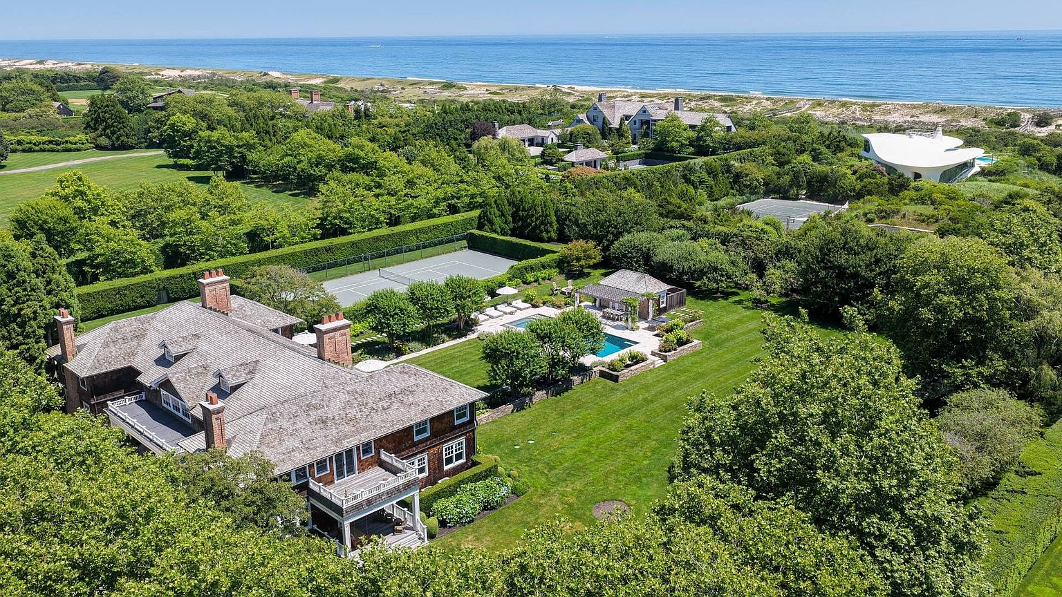 This expansive aerial view captures a luxurious coastal estate featuring a large, shingled residence with multiple chimneys and a wrap-around deck. The property includes a private tennis court, a swimming pool with a surrounding patio, and a separate pool house, all nestled within lush, manicured landscaping. The estate is situated near the ocean, offering a serene and private setting with a clear view of the coastline.