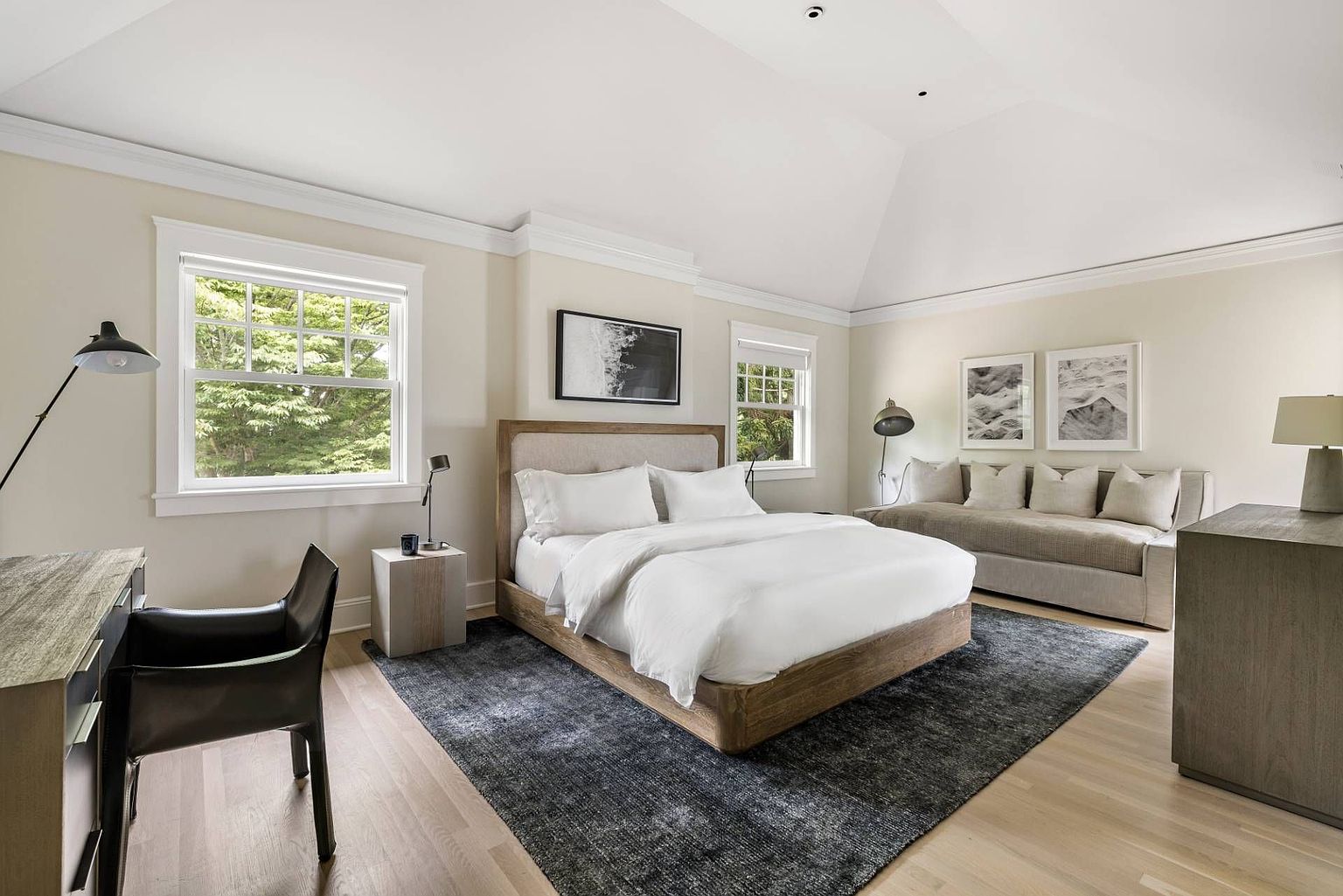 This spacious primary bedroom features a neutral color palette, high vaulted ceilings, and light wood flooring. The room is anchored by a large bed with a fabric headboard, flanked by minimalist side tables, and includes a dedicated seating area with a long bench against the wall. A desk setup is positioned to the left, while large windows allow for ample natural light, creating a serene and sophisticated atmosphere.