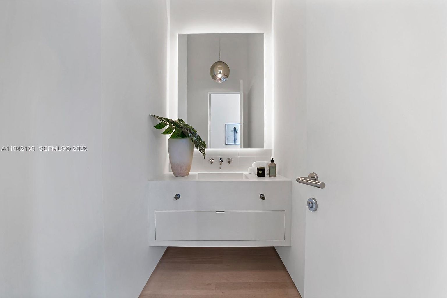 This minimalist powder room features a sleek, wall-mounted white vanity with a rectangular integrated sink and a large, backlit mirror. A decorative potted plant adds a touch of greenery to the clean, bright space, which is finished with light wood flooring. The perspective is a straight-on shot, highlighting the modern, uncluttered aesthetic and the soft, ambient glow of the integrated lighting.