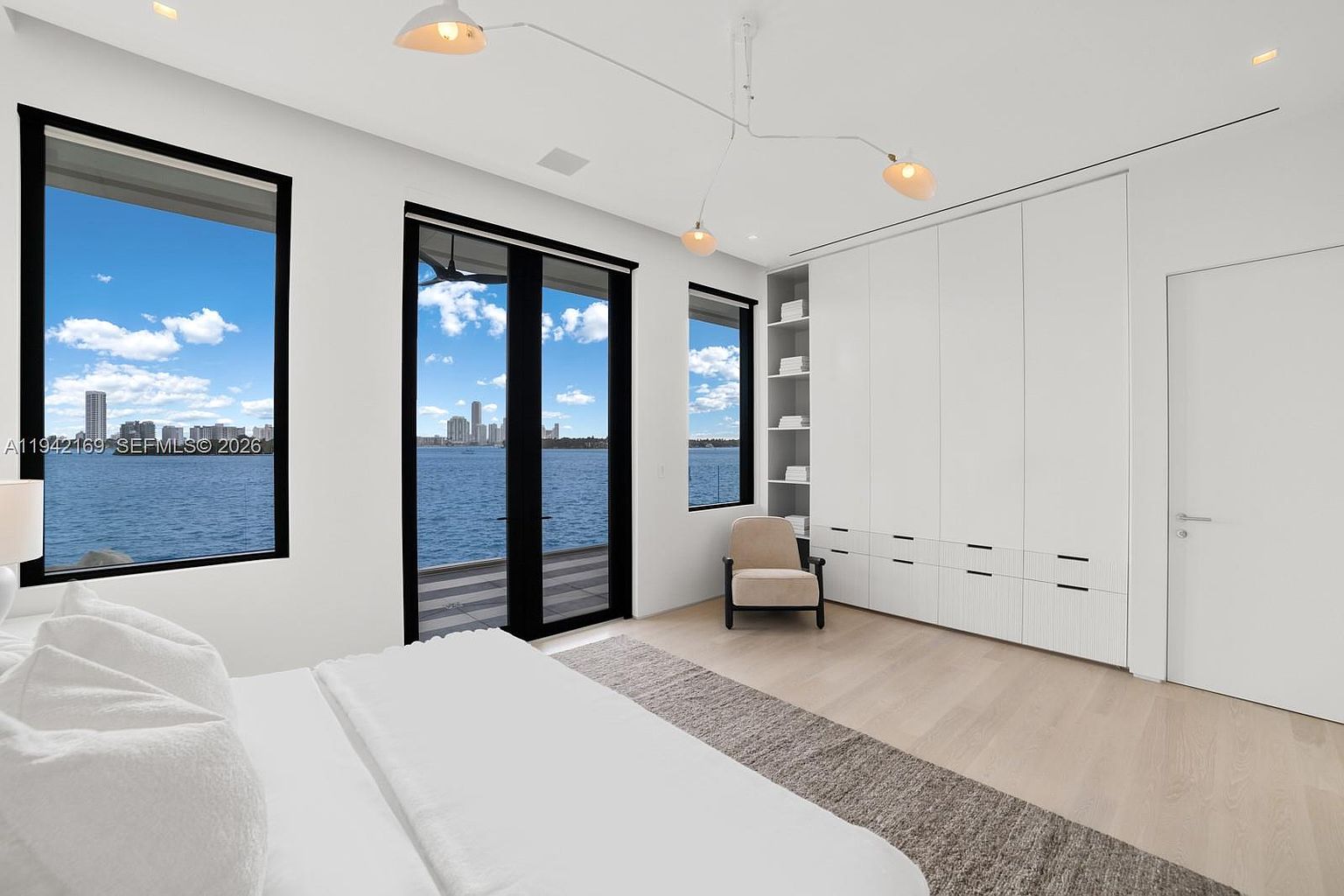 This bright, minimalist primary bedroom features a crisp white color palette and light wood flooring, creating an airy and serene atmosphere. Large windows and a glass door provide expansive, unobstructed views of the water and city skyline, while a built-in storage wall with integrated shelving offers clean, functional organization. A single neutral-toned armchair sits in the corner, complemented by modern, sculptural pendant lighting that adds a touch of contemporary elegance.