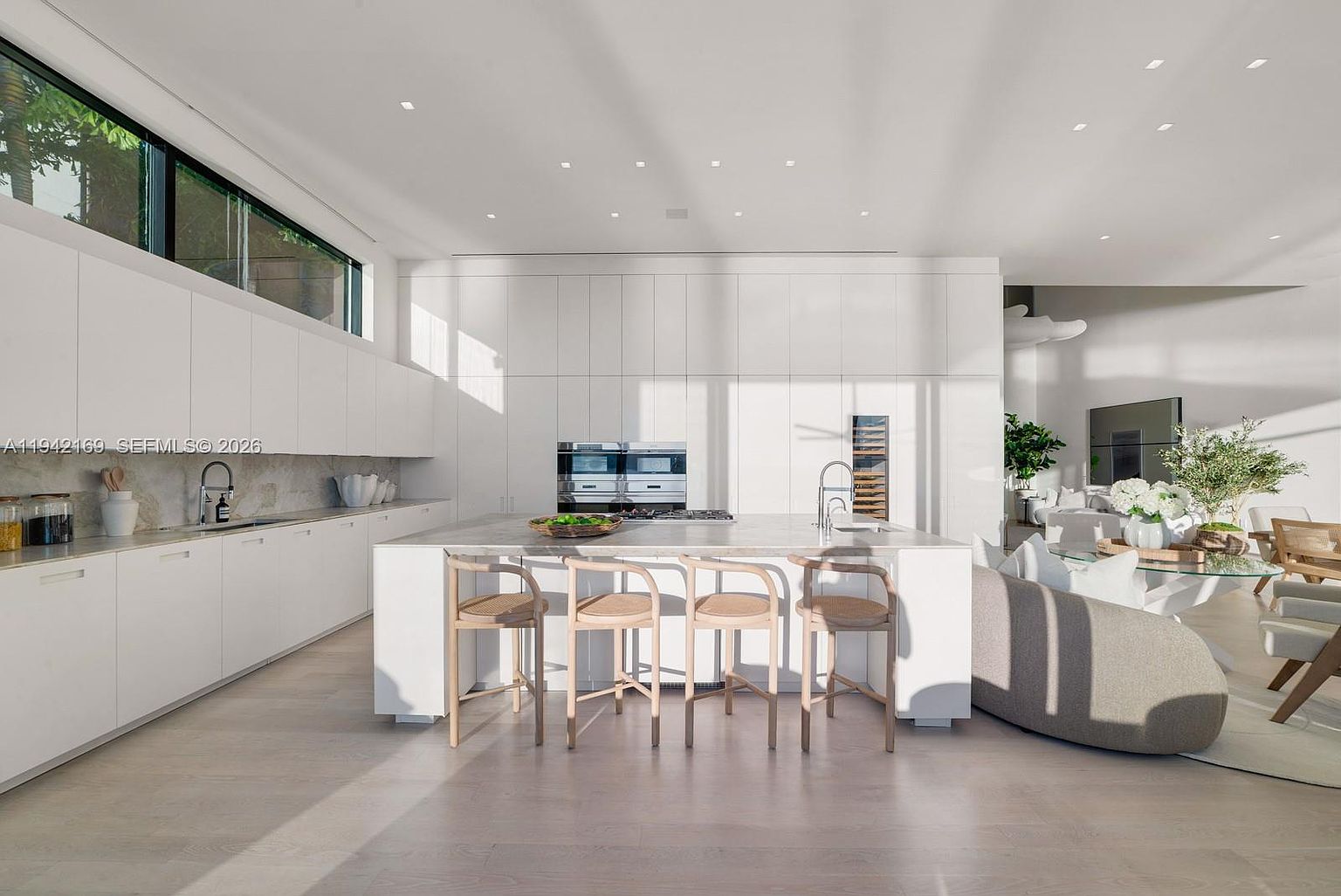 This bright, modern kitchen features sleek white cabinetry and a large central island with a stone countertop, providing ample workspace and seating. The space is illuminated by recessed lighting and a long, horizontal window that brings in natural light, while the open-concept design flows seamlessly into an adjacent living area. The minimalist aesthetic is complemented by light-toned wood flooring and high-end built-in appliances, creating a clean and sophisticated atmosphere.