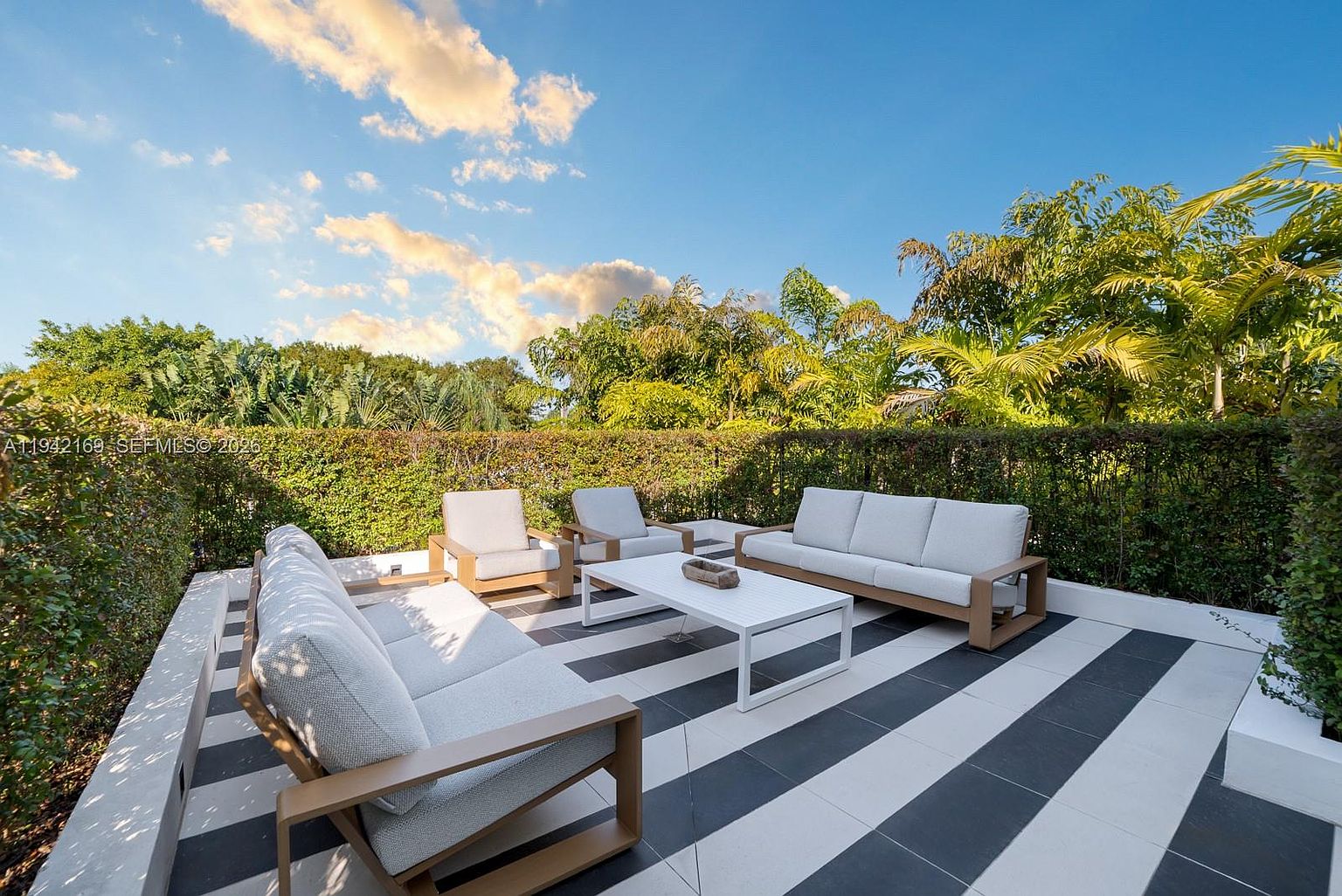 This outdoor patio features a striking black and white striped tile floor, creating a bold, modern aesthetic. The space is furnished with two light-toned sofas and two matching armchairs arranged around a central white coffee table, all set against a backdrop of lush green hedges and mature trees. The perspective is a wide-angle shot from a slightly elevated position, capturing the inviting and serene atmosphere of this private outdoor living area.