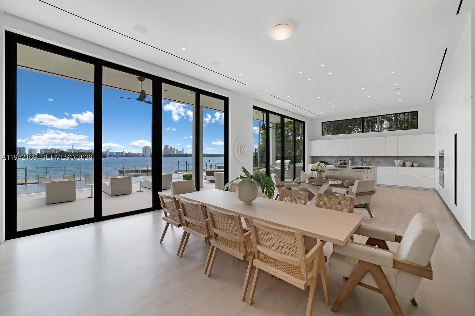 This open-concept living space features a long dining table with woven chairs in the foreground, transitioning into a bright kitchen area with a central island. The room is defined by expansive floor-to-ceiling glass walls that provide an unobstructed view of the waterfront and outdoor patio. The interior design is minimalist and airy, characterized by light wood flooring, white walls, and recessed lighting, creating a seamless connection between the indoor living area and the scenic exterior.