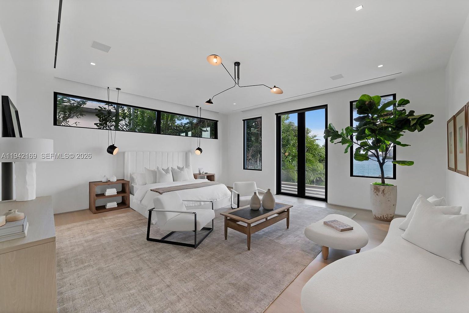 This spacious primary bedroom features a minimalist, contemporary design with a neutral color palette and light wood flooring. The room is anchored by a large bed with a tall, tufted headboard, flanked by wooden nightstands and modern pendant lighting. Large windows and glass doors provide abundant natural light and views of the outdoors, while a seating area with a curved sofa and a potted plant adds a touch of comfort and elegance.
