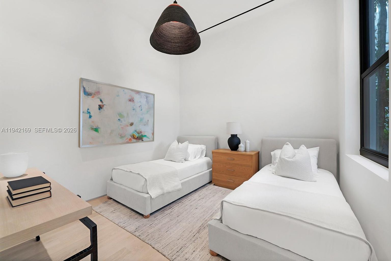 This bright and airy guest bedroom features two twin beds with light-colored upholstered headboards, positioned on either side of a wooden nightstand. A large, abstract piece of art hangs on the wall above one bed, while a modern, dark pendant light fixture hangs from the high ceiling. The room is finished with light wood flooring, a textured area rug, and a large window that allows for ample natural light.