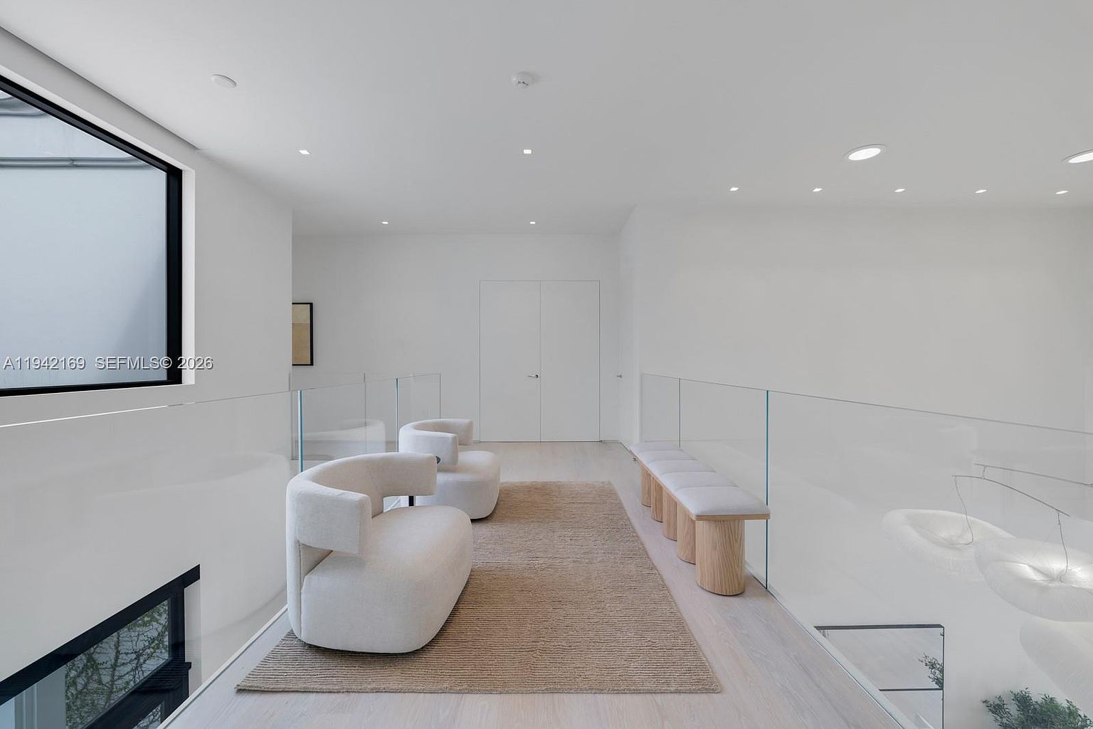 This bright, minimalist landing area features a sleek glass railing overlooking a staircase and a neutral-toned area rug centered between two curved, modern lounge chairs. To the right, a long bench with light wood legs provides additional seating, while the clean white walls and light-toned flooring create an airy, gallery-like atmosphere. The perspective is captured from the top of the stairs, looking across the open mezzanine toward a set of closed double doors.