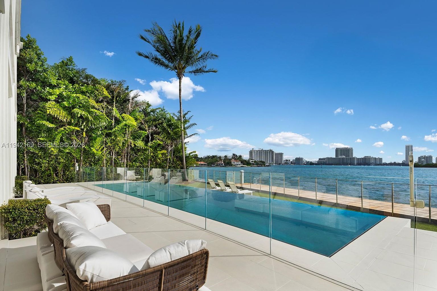 This luxurious outdoor living space features a sleek, rectangular infinity-edge pool that seamlessly blends into the waterfront view. A spacious patio area is furnished with comfortable, modern lounge seating, while a glass railing provides an unobstructed view of the bay and distant city skyline. The scene is framed by lush tropical greenery and a tall palm tree, creating a serene and high-end resort-like atmosphere.
