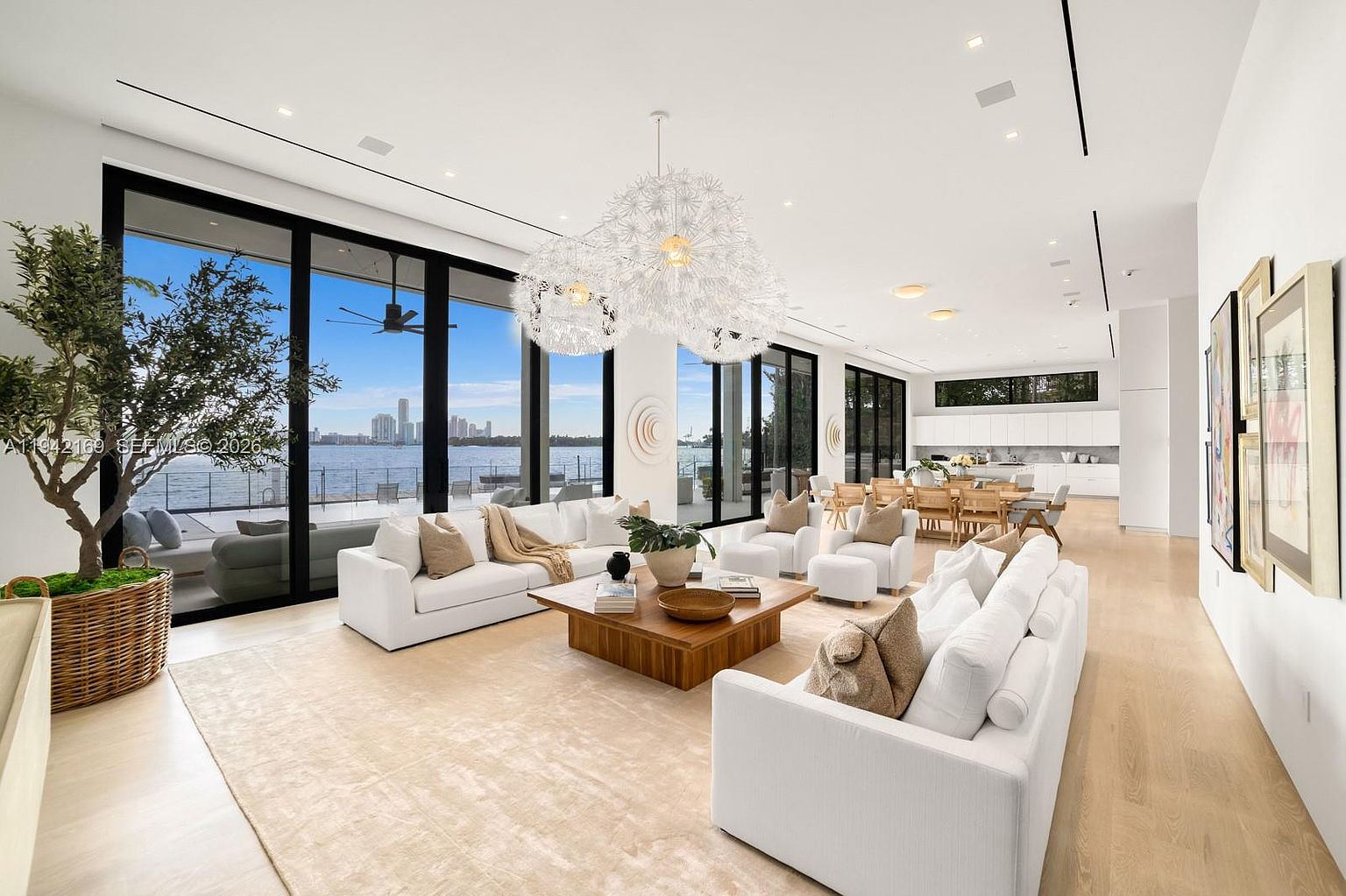 This expansive, open-concept living area features a bright, modern aesthetic with light wood flooring and a neutral color palette. The space is anchored by two large white sofas facing a central wooden coffee table, illuminated by a striking, sculptural dandelion-style chandelier. Large glass doors provide an unobstructed view of the waterfront, seamlessly blending the indoor living space with the exterior scenery.