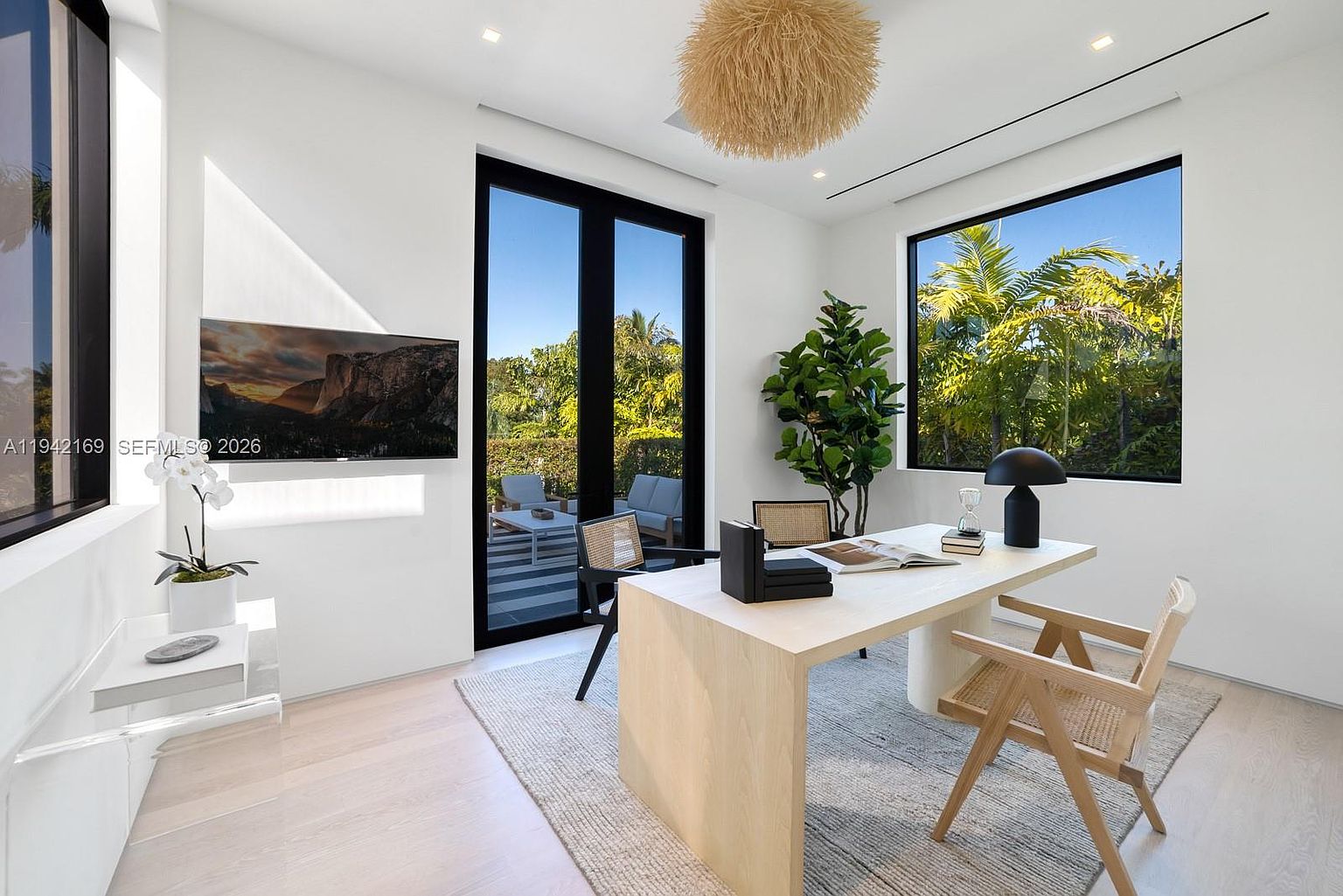 This bright and modern home office features a minimalist light wood desk positioned on a textured area rug. The room is accented by a large, textured pendant light fixture, a tall potted plant, and a wall-mounted television, with large windows and glass doors providing ample natural light and views of the outdoor patio. The clean white walls and neutral color palette create a professional yet serene workspace.
