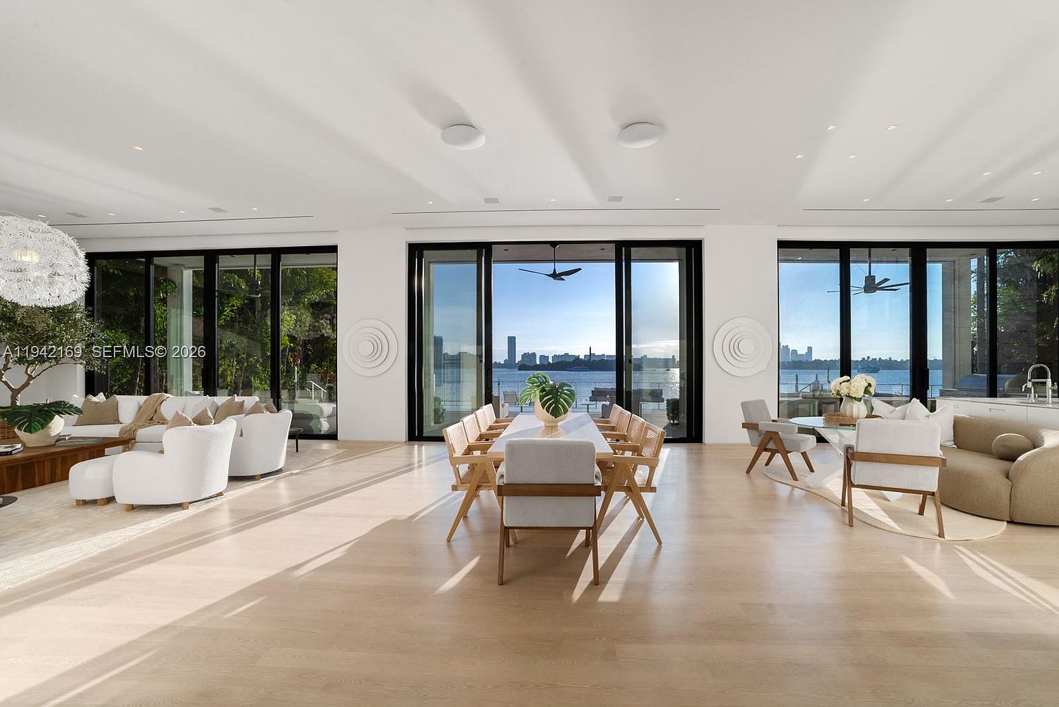 This expansive, open-concept living and dining area features light wood flooring and a neutral color palette that emphasizes the bright, airy atmosphere. Large floor-to-ceiling windows provide panoramic views of the water and city skyline, seamlessly connecting the interior space with the outdoors. The room is furnished with contemporary, curved seating and a long dining table, creating a sophisticated and inviting space for entertaining.