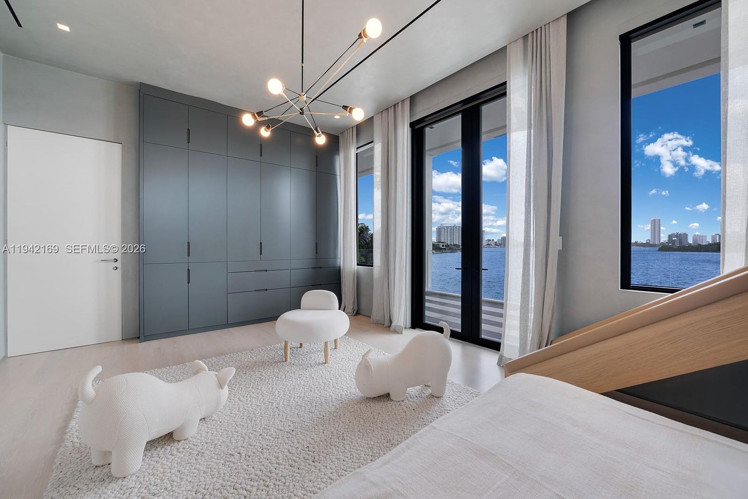 This modern bedroom features a minimalist aesthetic with light wood flooring, a textured area rug, and a large wall of dark grey cabinetry for storage. A unique, multi-armed chandelier hangs from the ceiling, while floor-to-ceiling windows and a glass door provide expansive views of the water and city skyline. The room is accented with playful, sculptural animal-shaped stools, creating a sophisticated yet whimsical atmosphere.