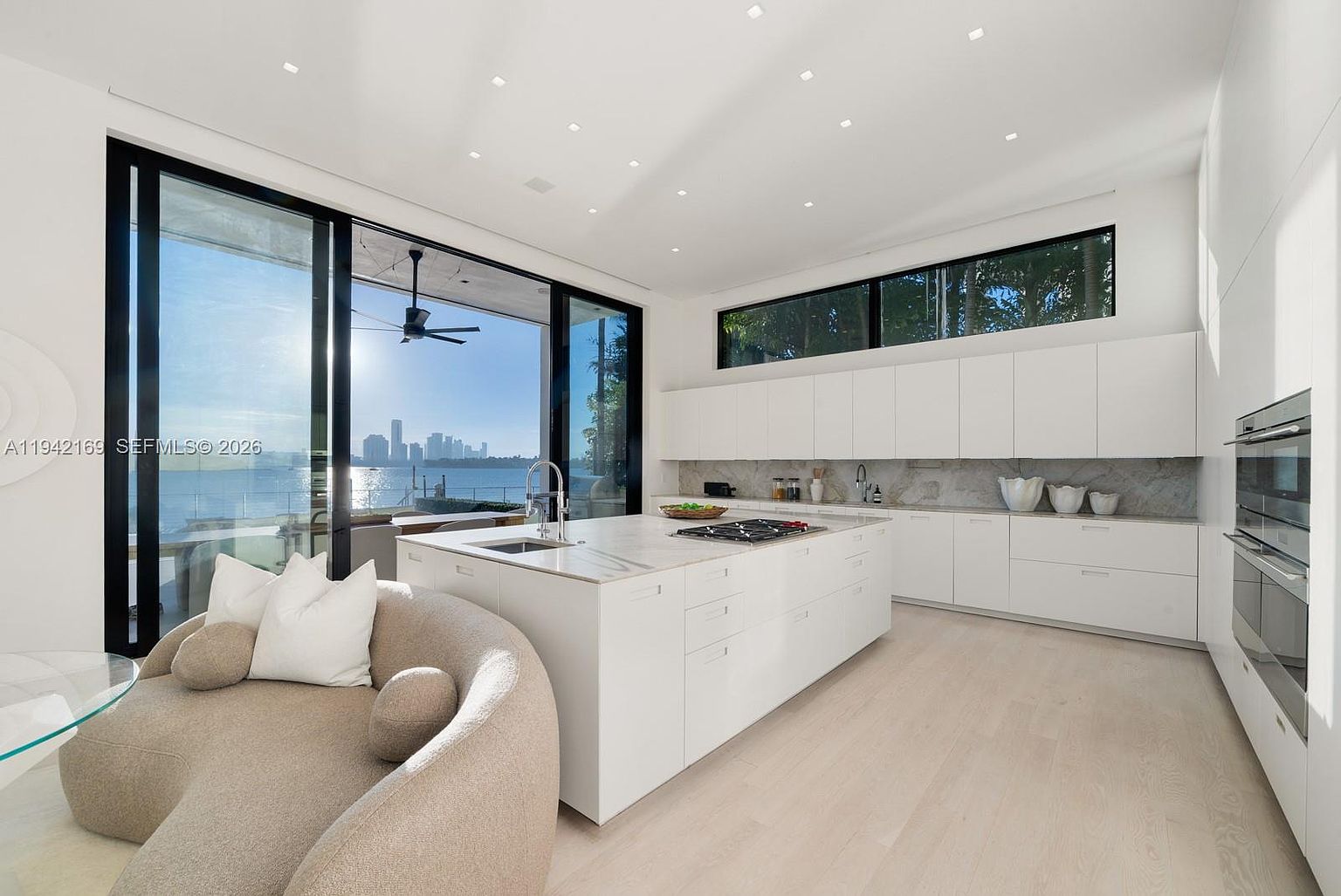 This modern, minimalist kitchen features sleek white cabinetry, a large central island with a stone countertop, and integrated appliances. The space is defined by expansive floor-to-ceiling glass sliding doors that offer a panoramic view of the water and city skyline. Light-toned wood flooring and recessed ceiling lighting create a bright, airy atmosphere, while a curved, neutral-toned sofa in the foreground adds a touch of soft texture to the clean-lined design.