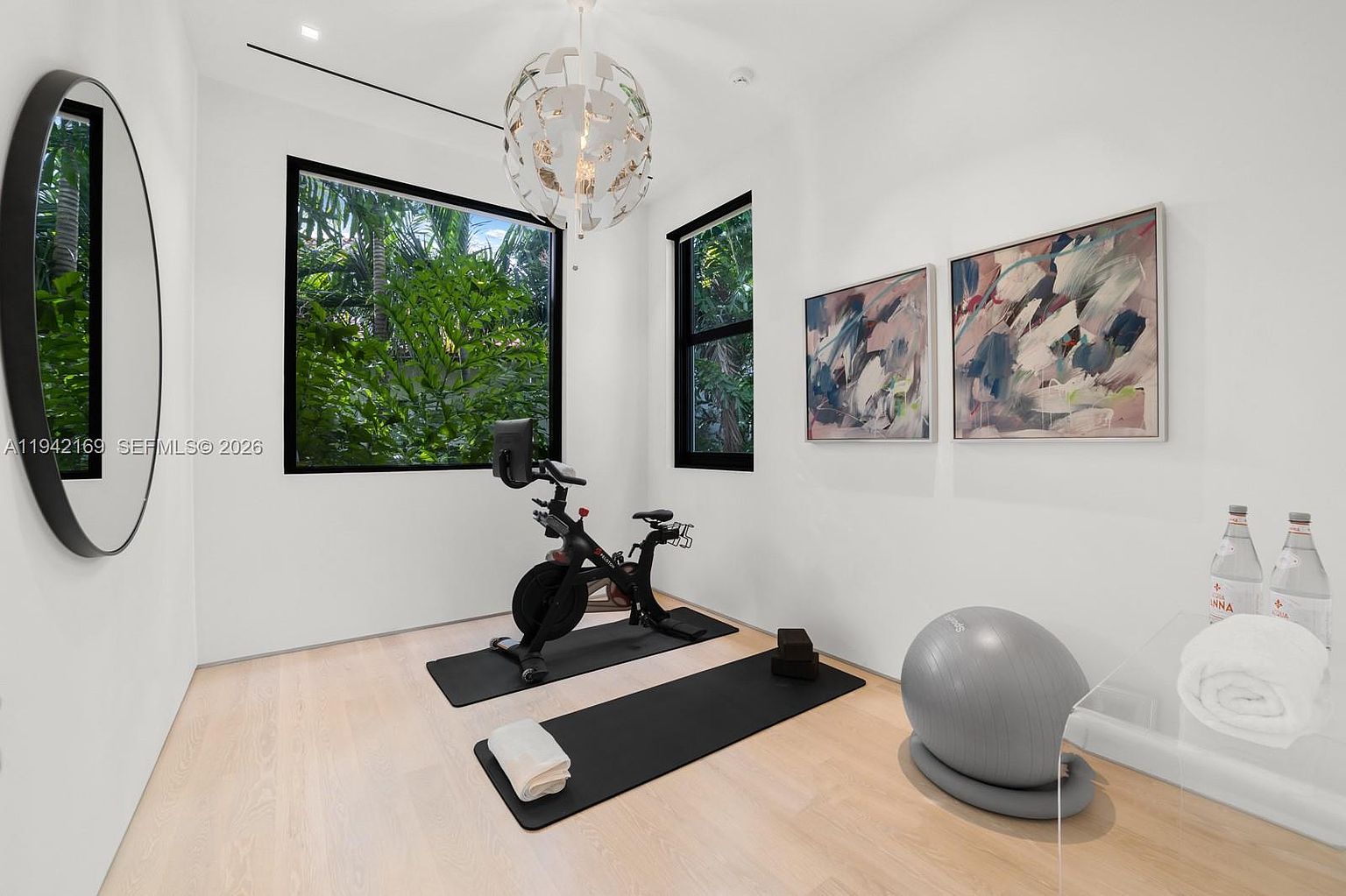This bright and modern home gym features light wood flooring and crisp white walls, creating an airy and inviting workout space. The room is equipped with a stationary exercise bike on a rubber mat, a yoga ball, and yoga blocks, while large windows provide a lush, green view of the outdoors. A unique, spherical chandelier hangs from the ceiling, and two abstract art pieces add a touch of color to the minimalist aesthetic.