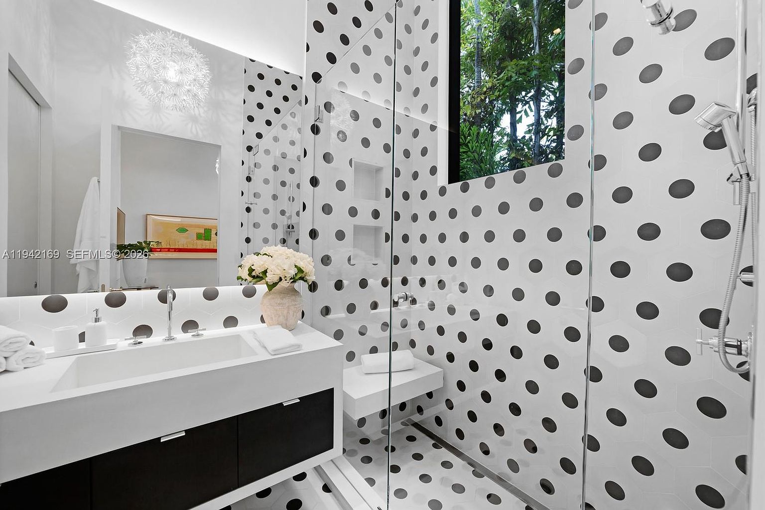 This modern bathroom features a striking, high-contrast aesthetic with white hexagonal wall and floor tiles adorned with large black polka dots. A sleek, floating vanity with a white countertop and dark cabinetry sits beneath a large mirror, complemented by a unique, textured spherical pendant light. A glass-enclosed shower area includes a built-in bench and a tall window that allows natural light to filter through, creating a bright and playful atmosphere.