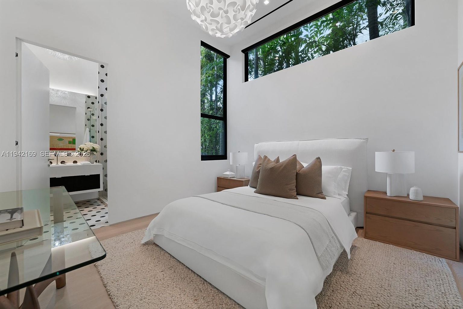 This bright and airy bedroom features a white upholstered bed frame centered against a wall with tall, narrow windows that provide a view of lush greenery. To the left, an open doorway reveals a bathroom with patterned tile flooring and a modern vanity, while a glass-topped desk sits in the foreground. The room is accented by a textured area rug, light wood nightstands, and a sculptural pendant light, creating a clean and contemporary aesthetic.