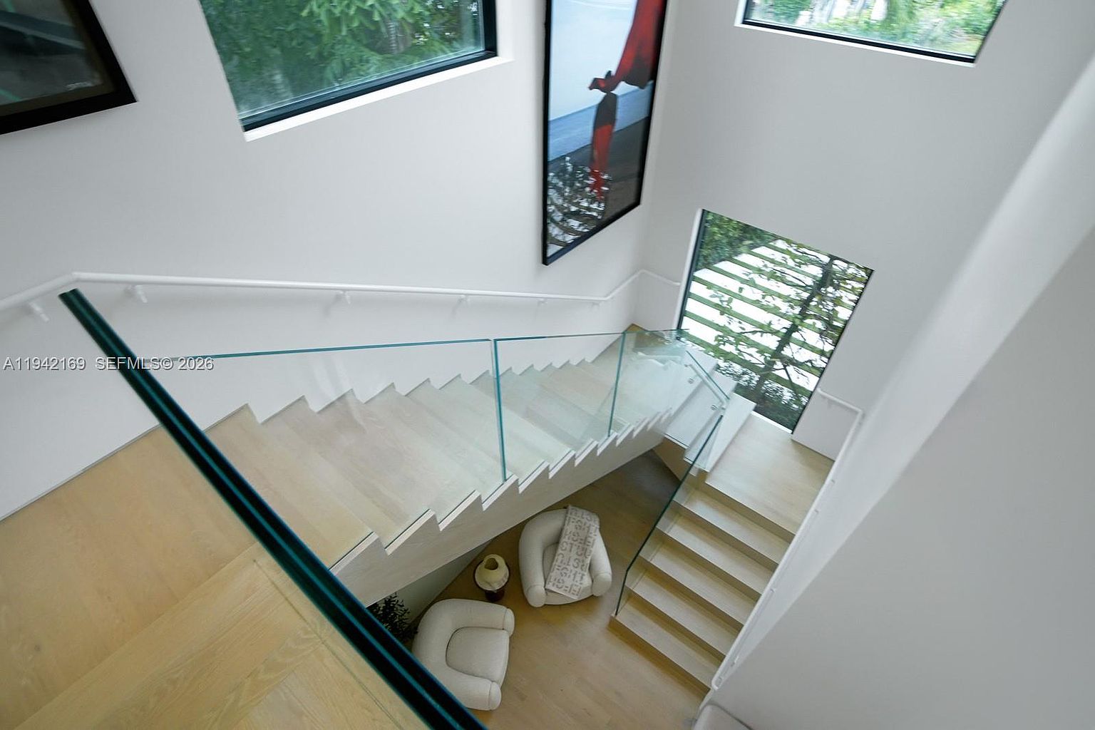 This high-angle shot captures a modern staircase featuring light wood treads and a sleek glass railing. The minimalist design is complemented by white walls and large windows that allow natural light to flood the space, highlighting a seating area with two white chairs at the base of the stairs. A tall, vertical piece of abstract art adds a bold pop of color to the otherwise neutral and airy architectural composition.