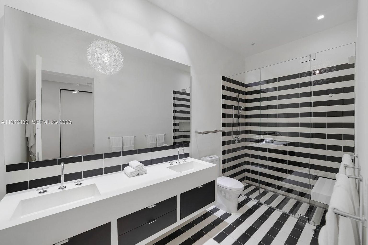 This modern bathroom features a striking high-contrast design with bold black and white horizontal stripes on the walls and floor. The space includes a long, dual-sink vanity with a white countertop, a large frameless glass-enclosed shower, and a contemporary toilet. A large mirror spans the width of the vanity, reflecting a unique dandelion-style pendant light fixture, creating a clean and dramatic aesthetic.