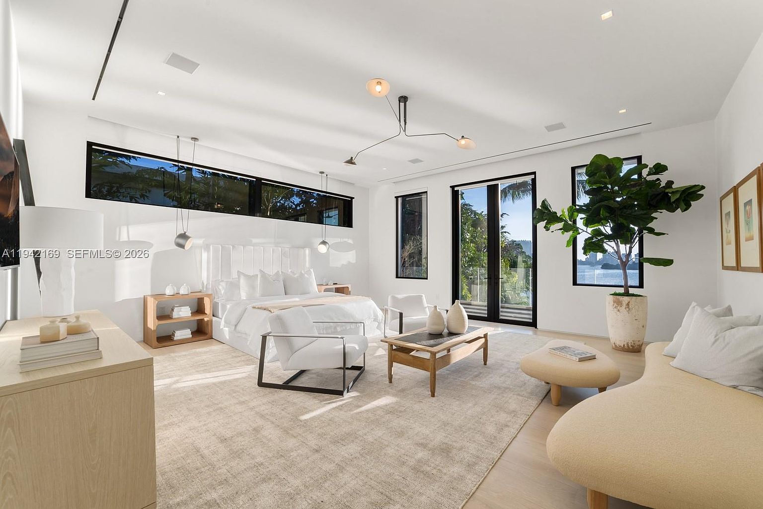This spacious primary bedroom features a minimalist, contemporary design with a neutral color palette and light wood flooring. The room is anchored by a large bed with a tall, tufted headboard, complemented by a seating area with a curved sofa and modern armchairs. Large windows and glass doors provide abundant natural light and views of the outdoors, while a sculptural chandelier adds a sophisticated focal point to the high ceiling.