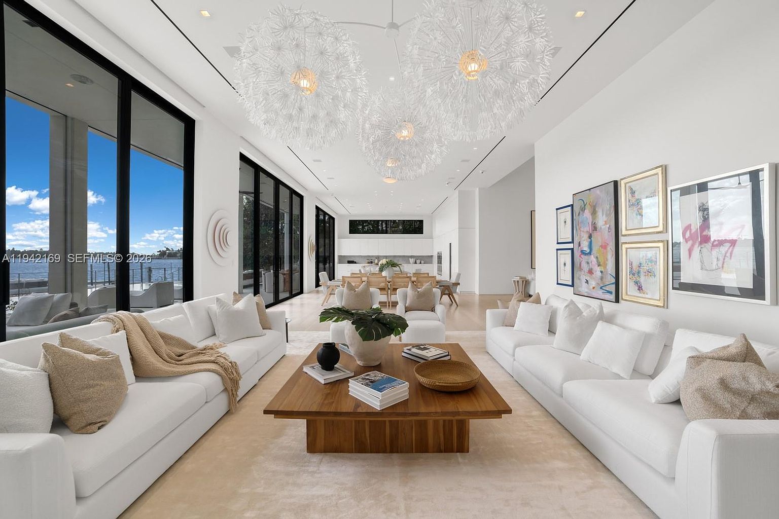 This expansive, open-concept living area features a bright, contemporary aesthetic with white walls and light-toned flooring. The space is anchored by two large white sofas facing a central wooden coffee table, illuminated by striking, oversized dandelion-style pendant lights. Large windows along the left wall provide an unobstructed view of the water, while the room flows seamlessly into a formal dining area in the background.