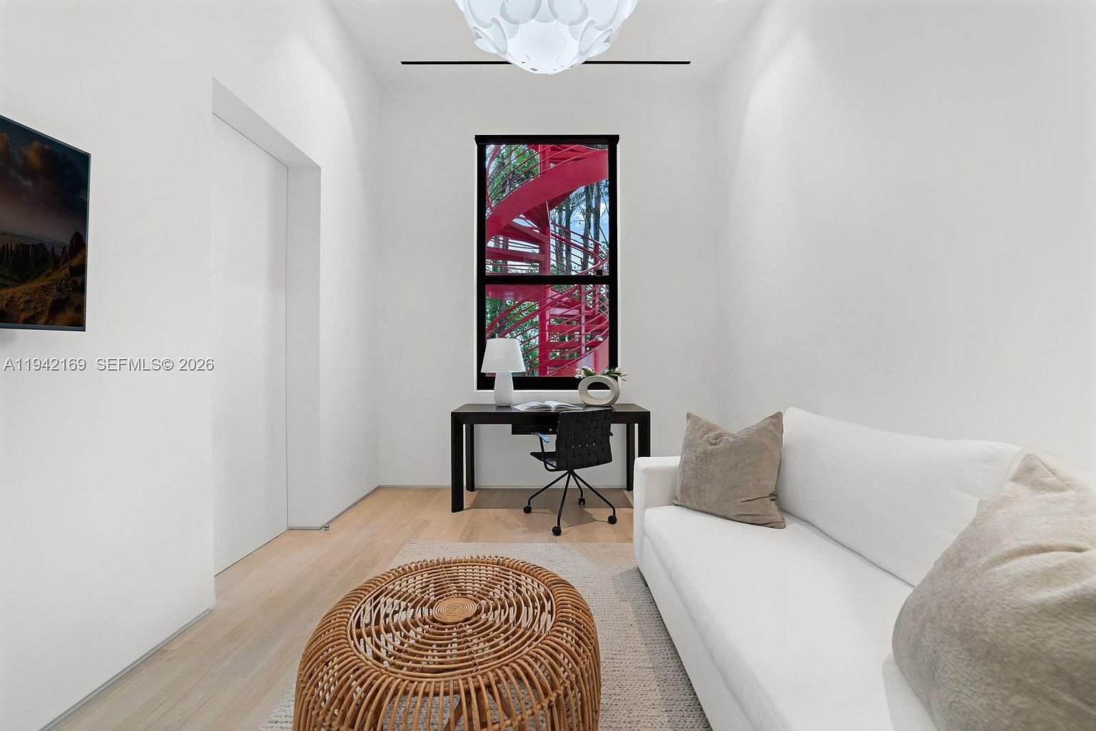 This bright, minimalist home office features a white sofa, a round woven coffee table, and a sleek desk positioned in front of a window. The window provides a unique view of a vibrant red spiral staircase outside, adding a bold pop of color to the otherwise neutral, light-filled space. A modern pendant light hangs from the ceiling, and the room is finished with light wood flooring and clean white walls.