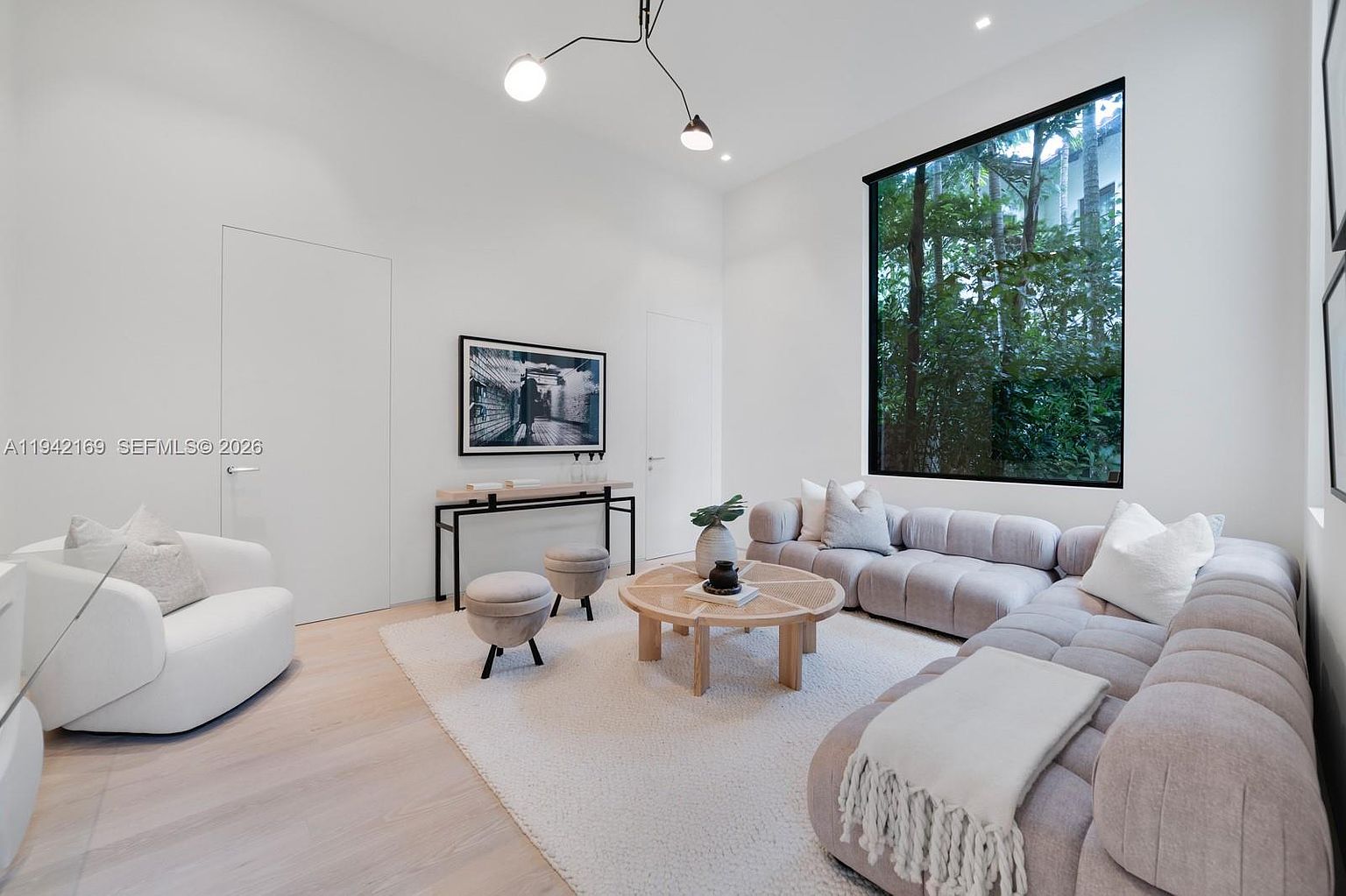 This bright and airy living room features a large, plush sectional sofa and a modern round coffee table centered on a textured area rug. A tall window provides a lush view of greenery, while a minimalist console table with two matching ottomans sits against the wall beneath a framed piece of art. The space is illuminated by a contemporary multi-arm light fixture, creating a sophisticated and inviting atmosphere.