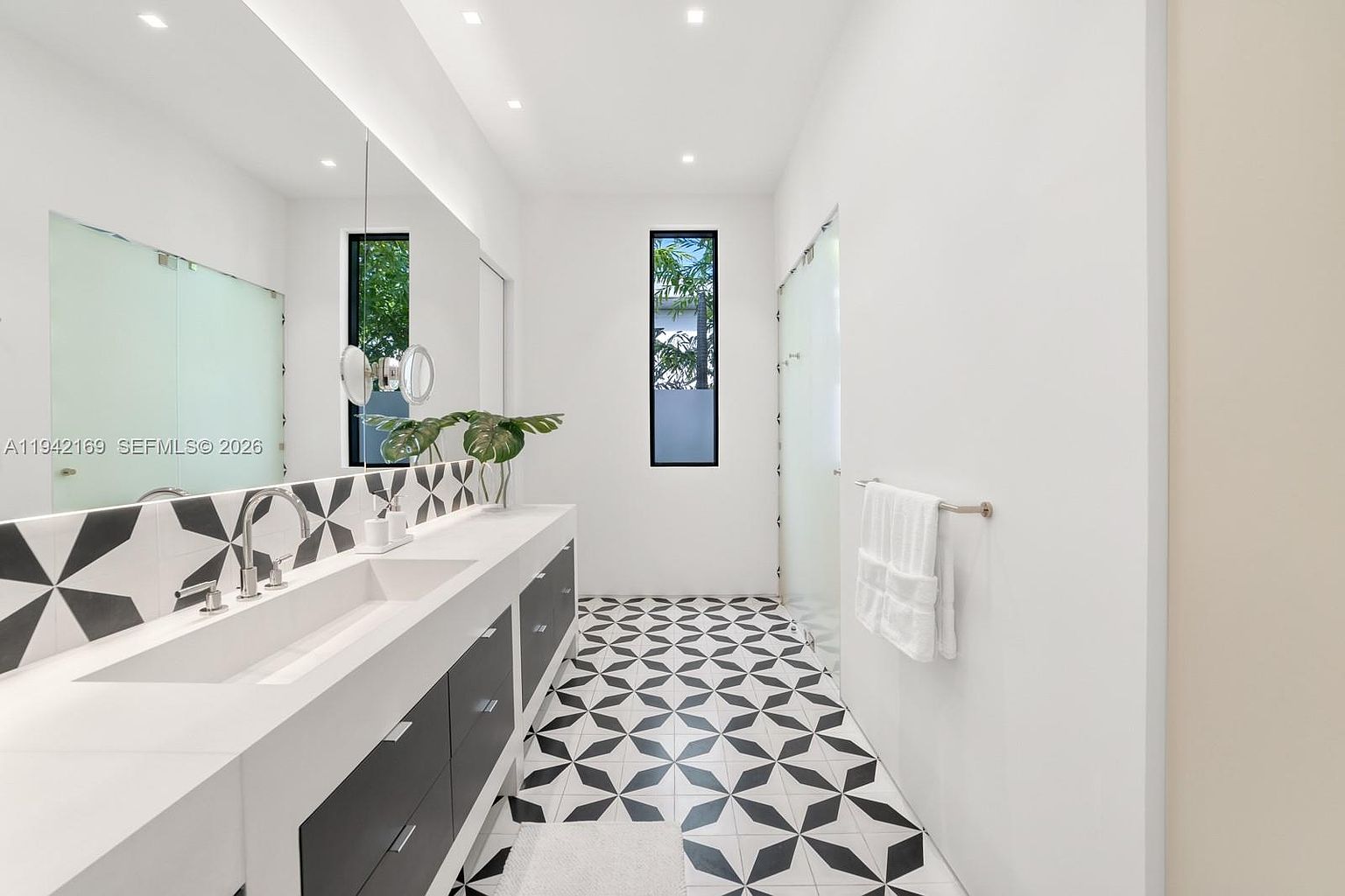 This modern bathroom features a long, floating vanity with a double sink and dark cabinetry, contrasted by a striking black-and-white geometric patterned tile floor. A large, frameless mirror spans the length of the vanity, reflecting the bright, minimalist space and a tall, narrow window. The room is finished with crisp white walls, recessed lighting, and a glass-enclosed shower area, creating a clean and contemporary aesthetic.