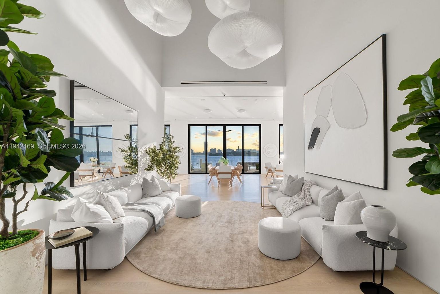 This expansive, high-ceilinged living room features a symmetrical layout with two plush white sofas facing each other across a large, circular area rug. The space is anchored by a massive piece of abstract wall art and a large mirror, while unique, cloud-like pendant lights hang from the vaulted ceiling. Through the rear, large glass doors provide a clear view of the water, creating a bright, airy, and sophisticated atmosphere.