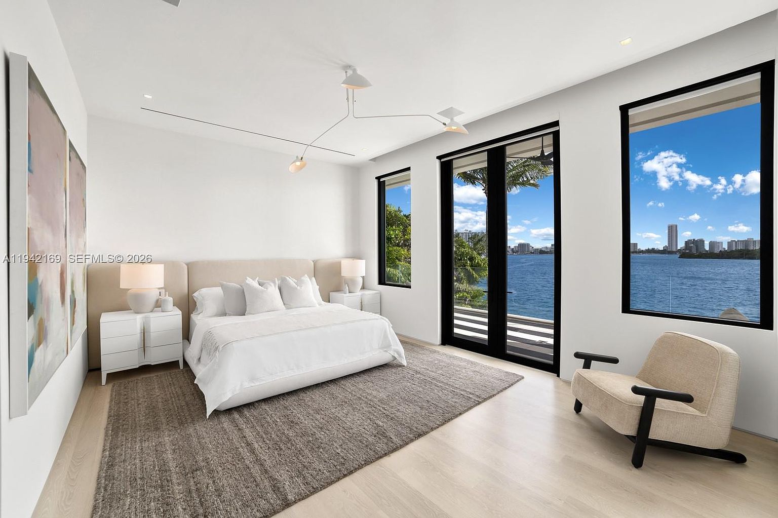 This spacious primary bedroom features a minimalist aesthetic with light wood flooring and a large, textured area rug anchoring the bed. The room is highlighted by expansive windows and glass doors that provide a panoramic view of the water and distant skyline. A modern, sculptural light fixture hangs from the ceiling, complementing the clean lines of the neutral-toned furniture and artwork.