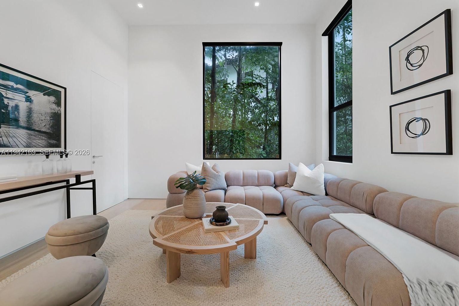 This bright and airy living room features a large, neutral-toned sectional sofa arranged around a circular, light wood coffee table with a woven top. A tall, rectangular window provides a clear view of lush greenery outside, while two framed abstract art pieces adorn the wall. The space is finished with light-colored flooring and a textured area rug, creating a clean, modern, and inviting atmosphere.