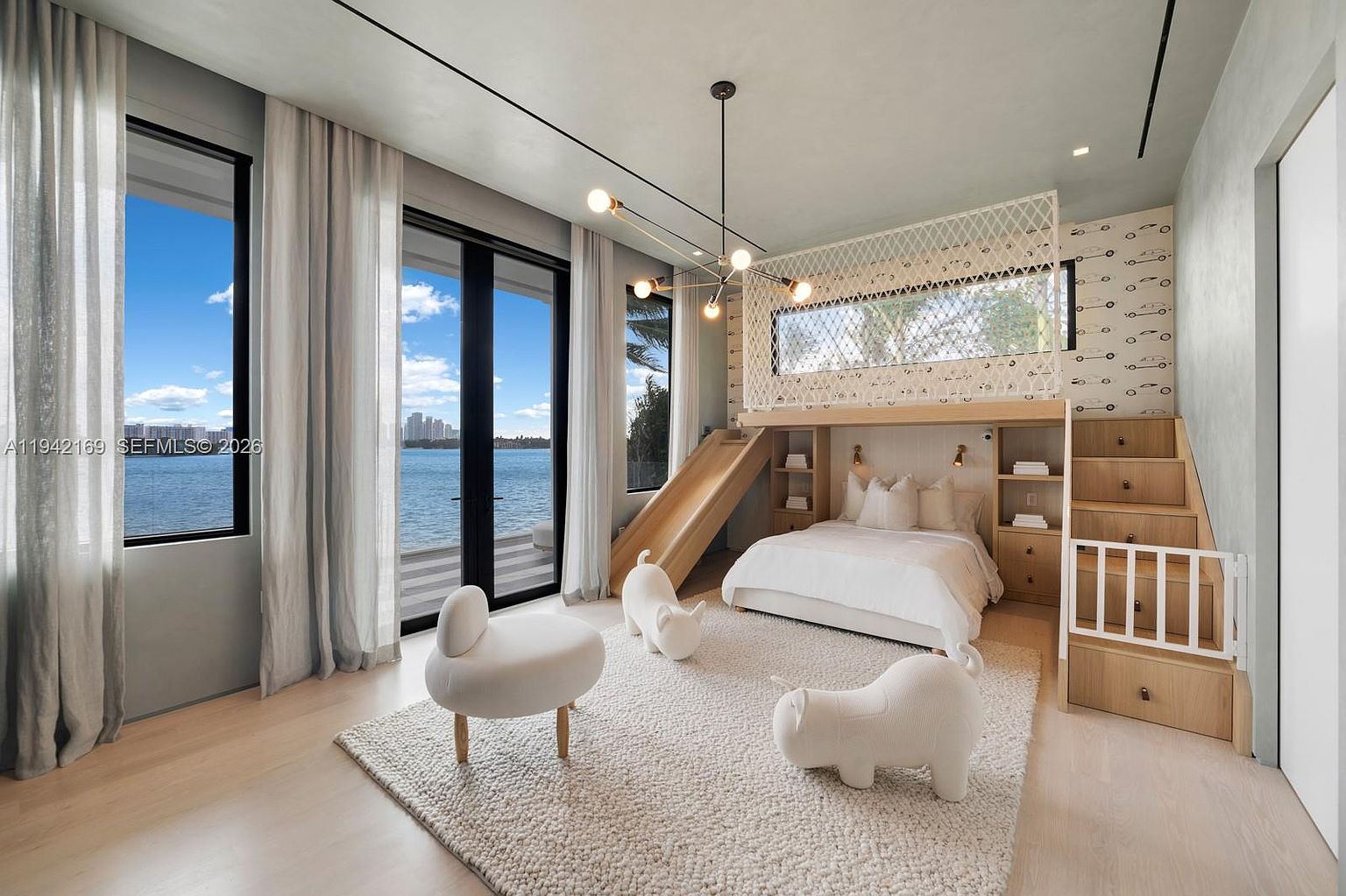 This whimsical and modern children's bedroom features a custom built-in bed structure that includes a slide and a staircase with a safety gate. The room is bright and airy, highlighted by large windows that offer a stunning, unobstructed view of the water and distant city skyline. A textured area rug sits beneath three playful, animal-shaped ottomans, creating a cozy and imaginative space.