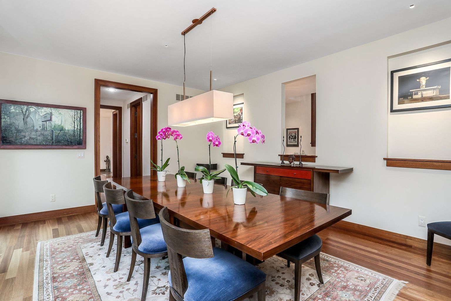 This is a well-appointed dining room featuring a long, polished wooden table set with floral arrangements. The room is furnished with blue upholstered chairs and a patterned rug, complemented by artwork and a modern rectangular chandelier. The space exudes a sense of elegance and comfort, ideal for formal dining and entertaining.