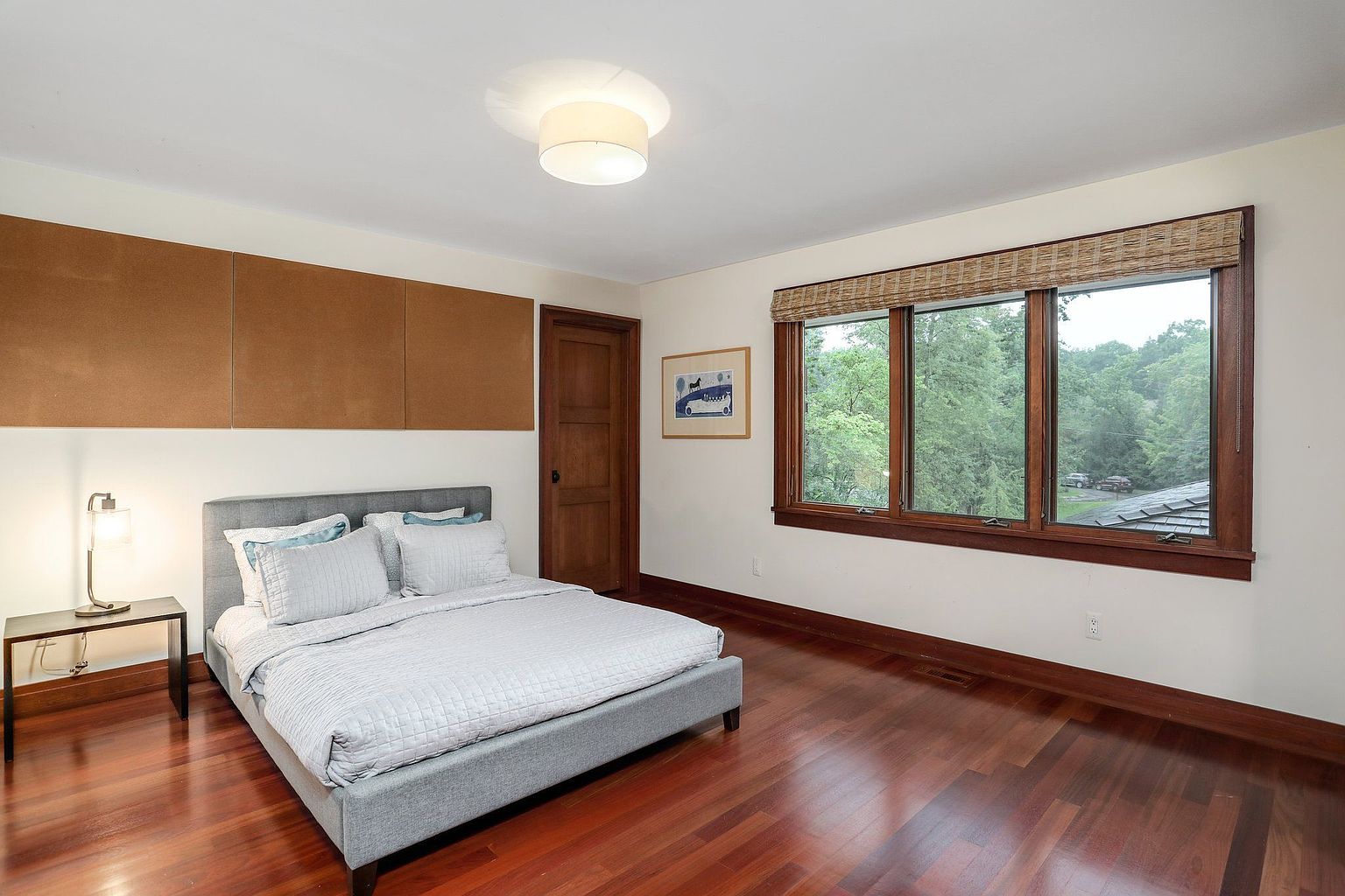 This is a well-lit bedroom featuring a gray upholstered bed with light blue accents, a dark wood nightstand with a lamp, and a large window offering a view of the outdoors. The room has hardwood floors and a neutral color palette, creating a calm and inviting atmosphere. A corkboard is mounted on the wall above the bed.