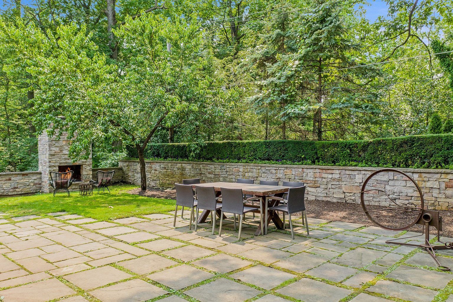 This image showcases a beautifully landscaped backyard featuring a stone patio with a dining table and chairs, perfect for outdoor entertaining. A stone fireplace and seating area add a cozy touch, while a decorative metal sculpture provides a unique focal point. The lush greenery and mature trees create a private and serene atmosphere.
