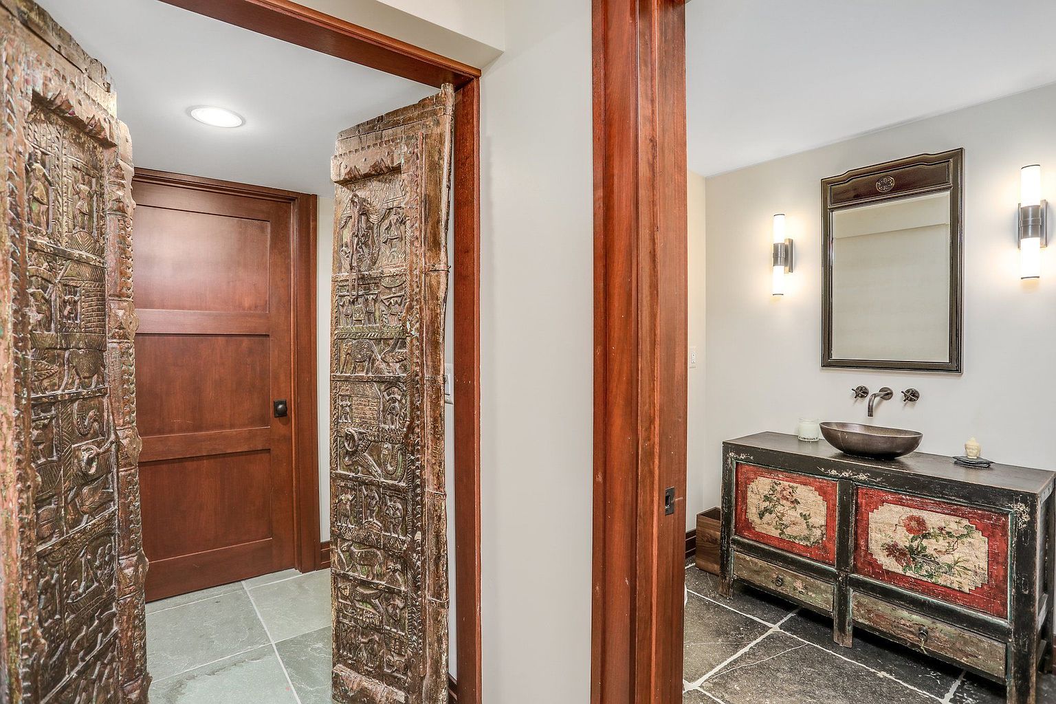 This image showcases a unique guest bathroom featuring an ornate vanity with a vessel sink and a decorative mirror. The walls are painted in a neutral tone, complemented by modern sconces. The flooring is dark tile, and an intricately carved wooden door adds an exotic touch to the space.