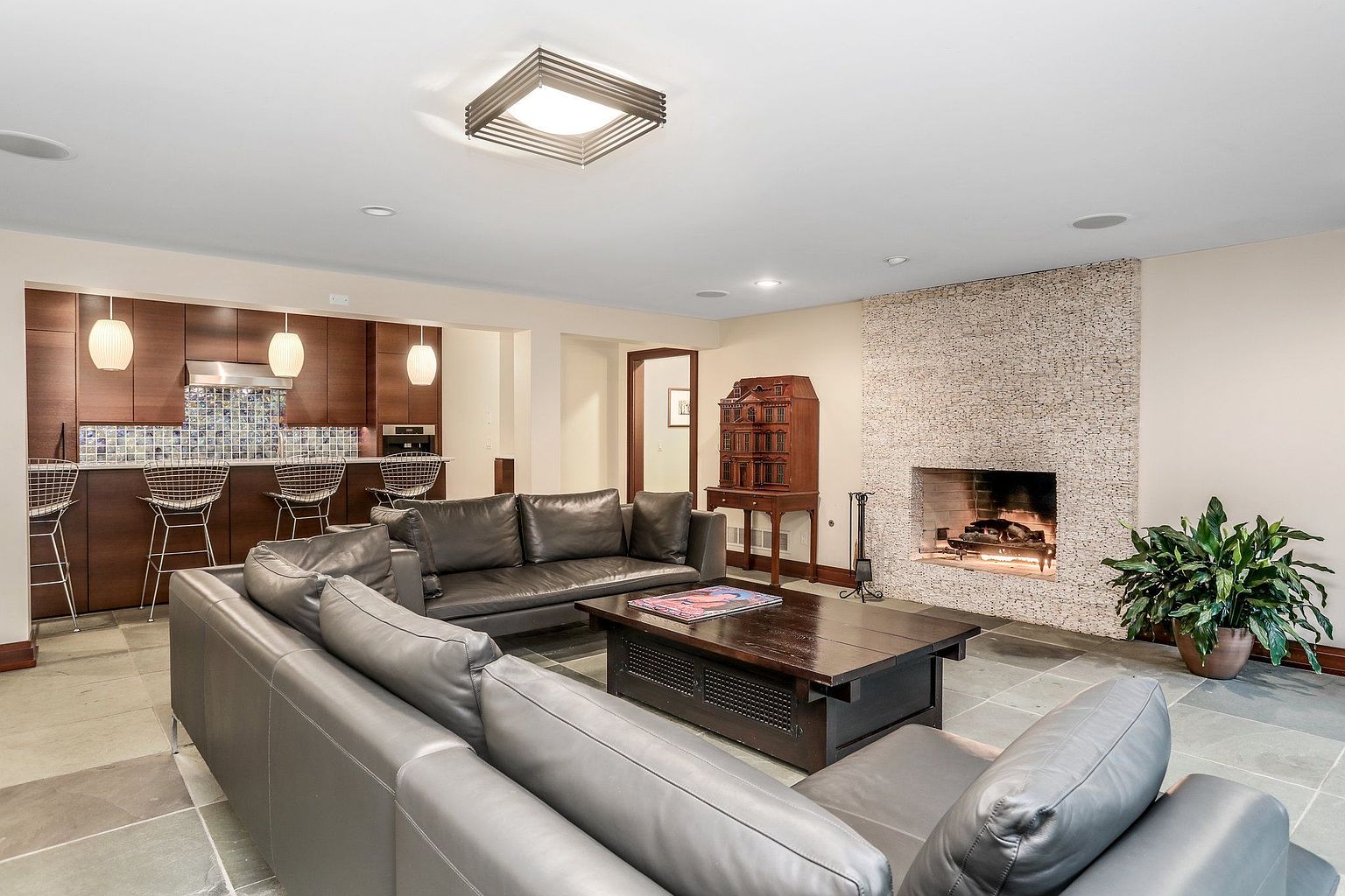 This is an interior shot of a living room featuring a modern design with a fireplace and a bar area. The room is furnished with a large gray leather sectional sofa and a dark wood coffee table. The fireplace is surrounded by a textured stone wall, and a plant sits beside it. The bar area has wooden cabinets, a tiled backsplash, and bar stools.