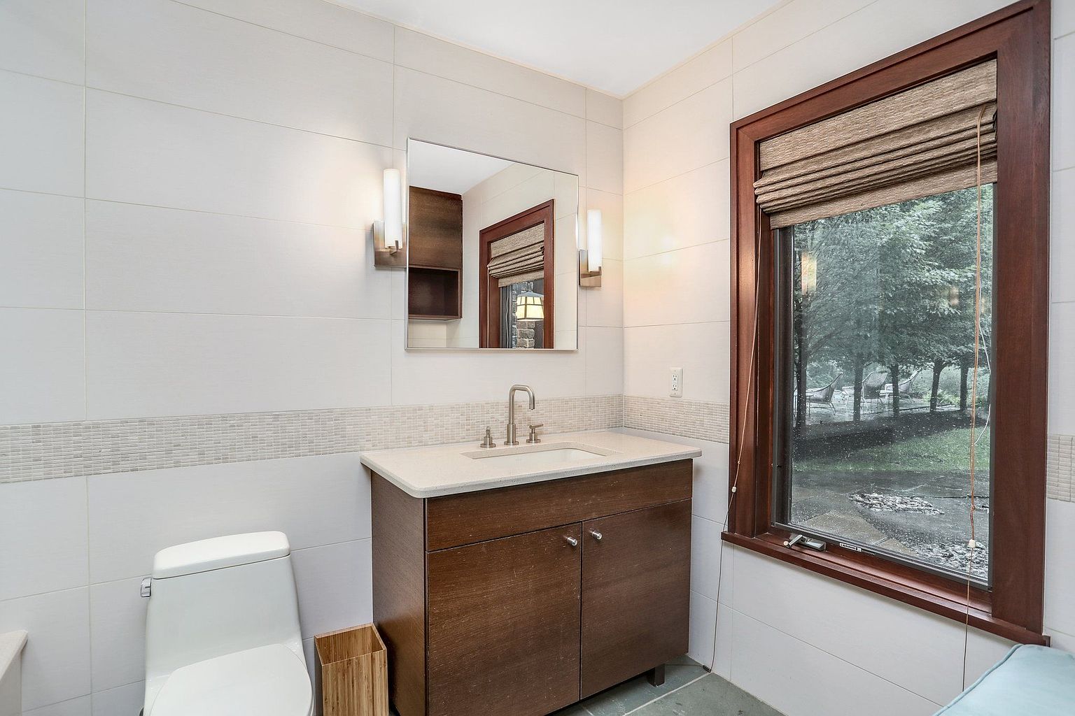 This is a well-lit bathroom featuring a modern vanity with a light countertop and dark wood cabinetry. A rectangular mirror hangs above the sink, flanked by sconce lighting. The walls are tiled in a neutral color, with a decorative mosaic tile strip adding visual interest. A toilet is visible to the left, and a window with a wooden frame and a woven shade offers a view of the outdoors.
