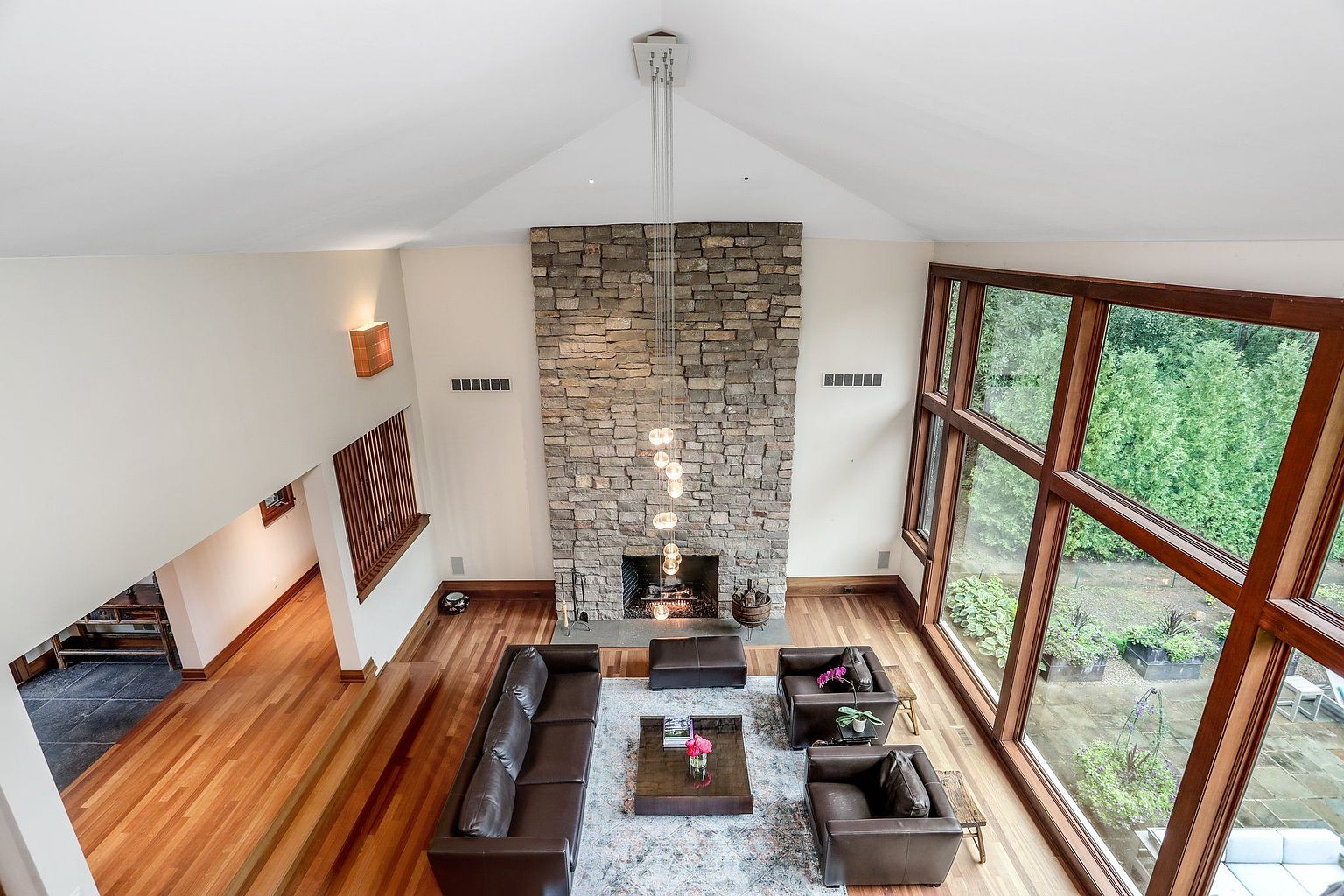 This is a high-angle shot of a spacious living room featuring a stone fireplace, a brown leather sofa and armchairs, and a large window offering natural light and views of the outdoors. The room has hardwood floors and a modern chandelier, creating a luxurious and inviting atmosphere. The vaulted ceiling adds to the sense of spaciousness.