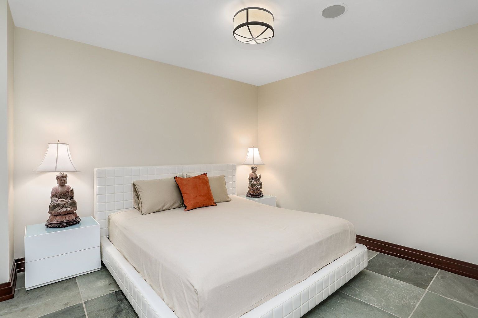 This is a well-lit bedroom featuring a white platform bed with a tiled headboard, complemented by two nightstands. Each nightstand is adorned with a lamp and a decorative Buddha statue. The room has a neutral color palette, with beige walls and a unique tiled floor, creating a serene and inviting atmosphere.
