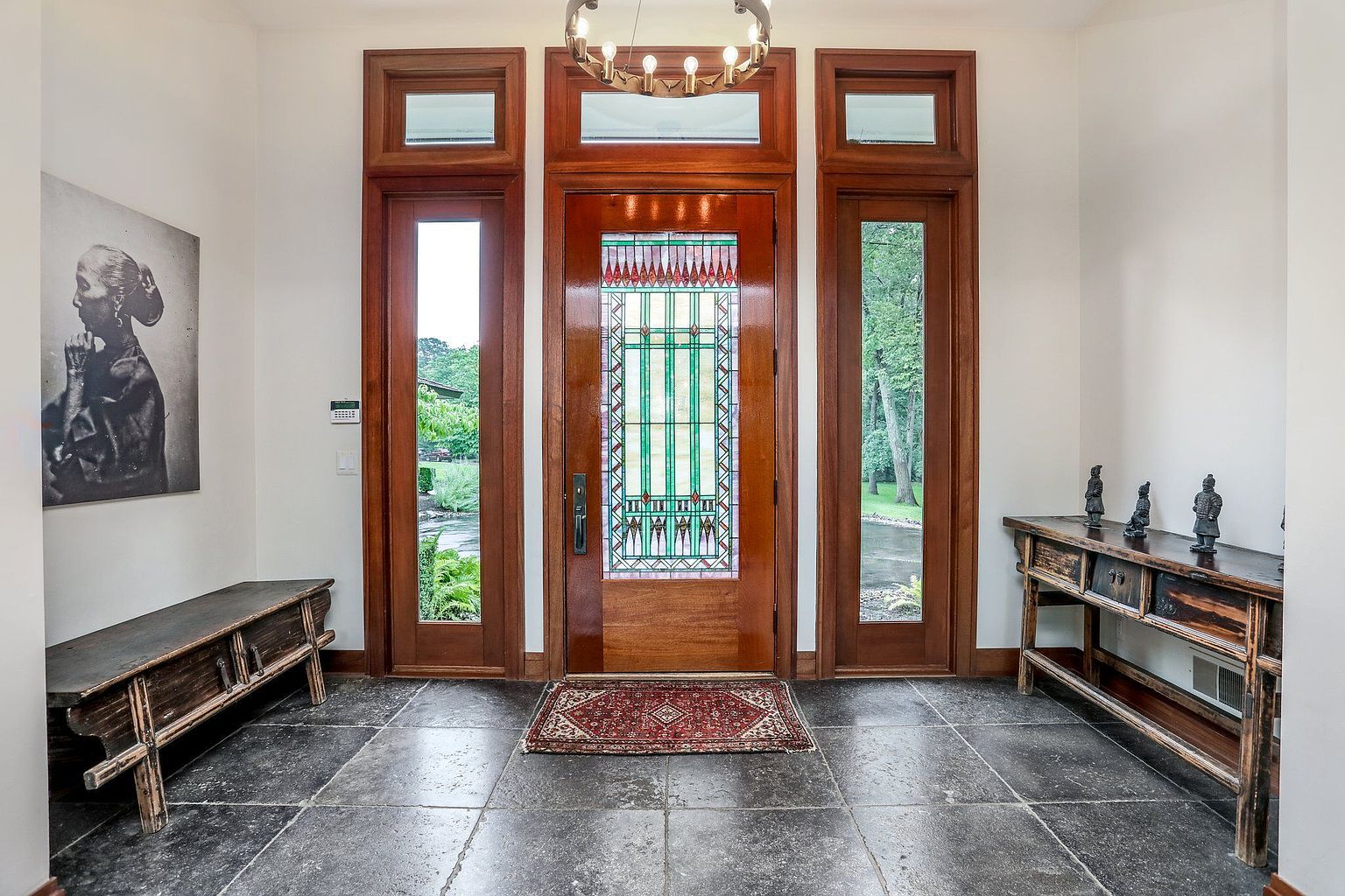 This is an interior shot of an entryway featuring a striking wooden door with stained glass, flanked by sidelights and topped with transom windows. The space is decorated with a dark wood bench on one side and a console table with decorative figurines on the other, set against a backdrop of neutral walls and dark stone flooring. A modern chandelier hangs from the ceiling, adding a touch of elegance.