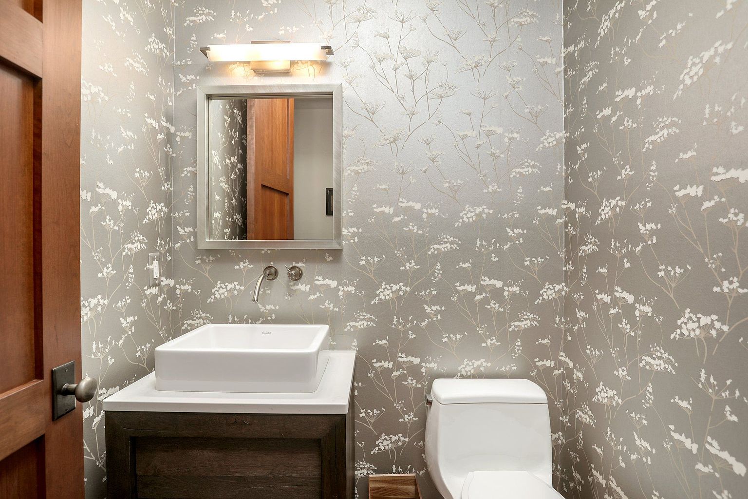 This is a well-appointed guest bathroom featuring a modern square sink atop a dark wood vanity with a white countertop. The walls are adorned with a delicate floral wallpaper, complemented by a sleek, square mirror and contemporary lighting fixture. A white toilet sits adjacent to the vanity, completing the space with a clean and stylish aesthetic.