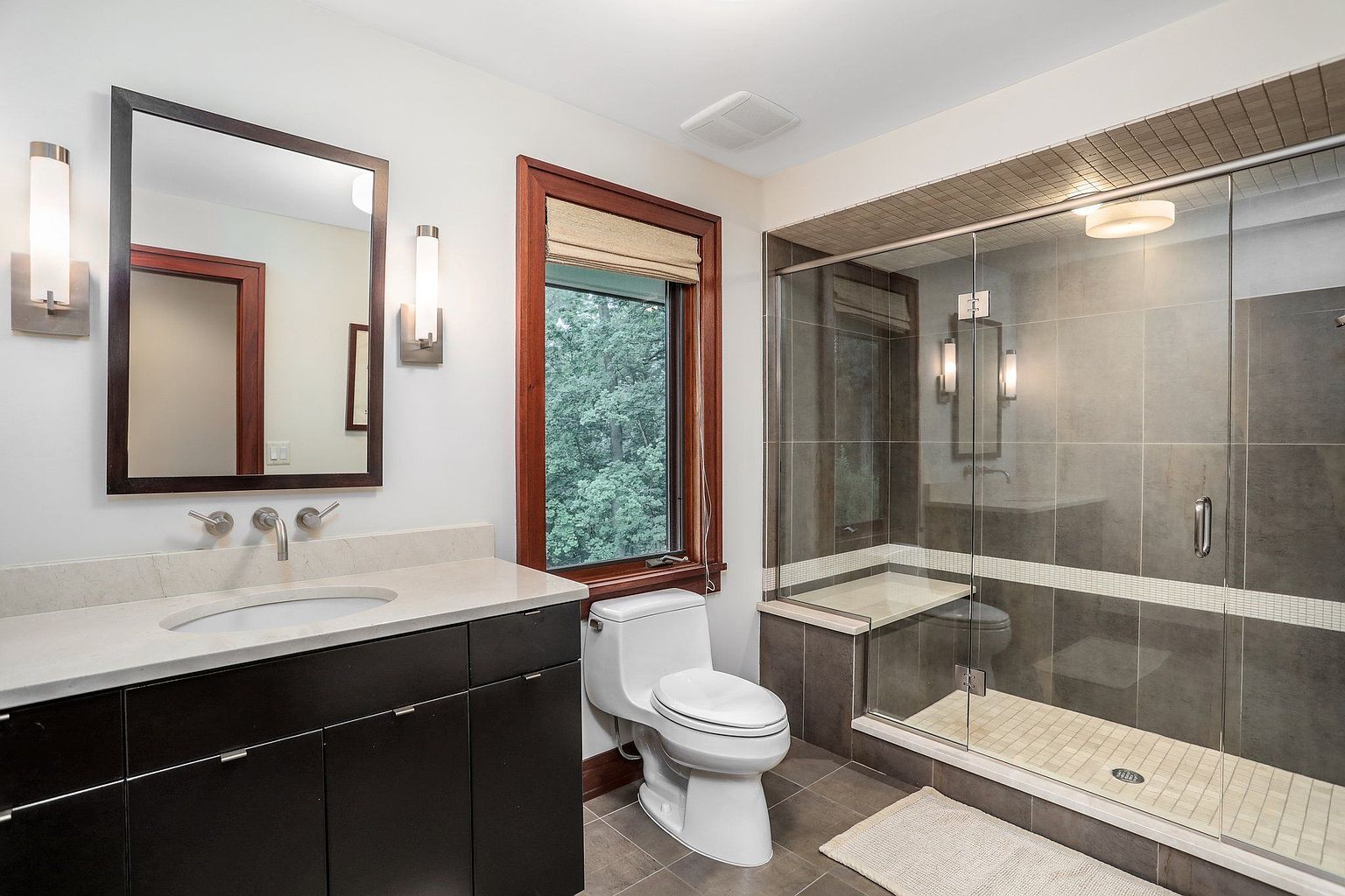 This is a well-lit primary bathroom featuring a dark wood vanity with a light countertop and a rectangular mirror. A modern toilet sits next to a window with a wooden frame, and a glass-enclosed shower with gray tiling and a built-in bench is visible. The overall impression is clean and contemporary.
