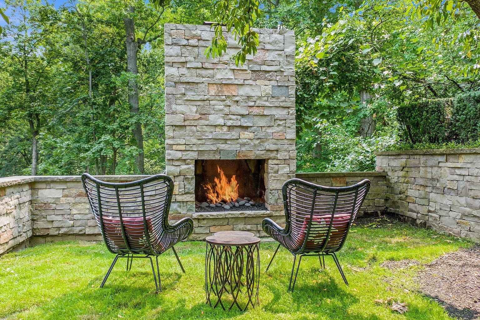 This image showcases a beautifully designed outdoor living space, featuring a stone fireplace as the focal point. Two modern chairs and a small table are arranged in front of the fireplace, creating an inviting seating area. The surrounding lush greenery and stone walls add to the serene and private atmosphere, perfect for relaxation and entertaining.
