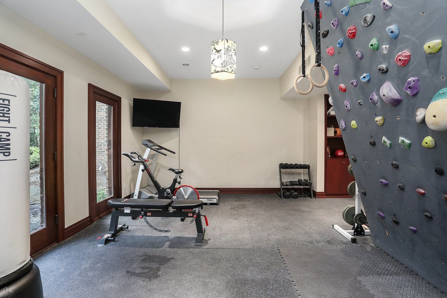 This is a well-equipped home gym featuring a climbing wall, exercise bike, treadmill, weight bench, and dumbbell rack. The room has a neutral color palette with dark wood trim around the doors and a gray rubber floor. The space is well-lit with recessed lighting and a decorative pendant light, creating a functional and inviting workout environment.