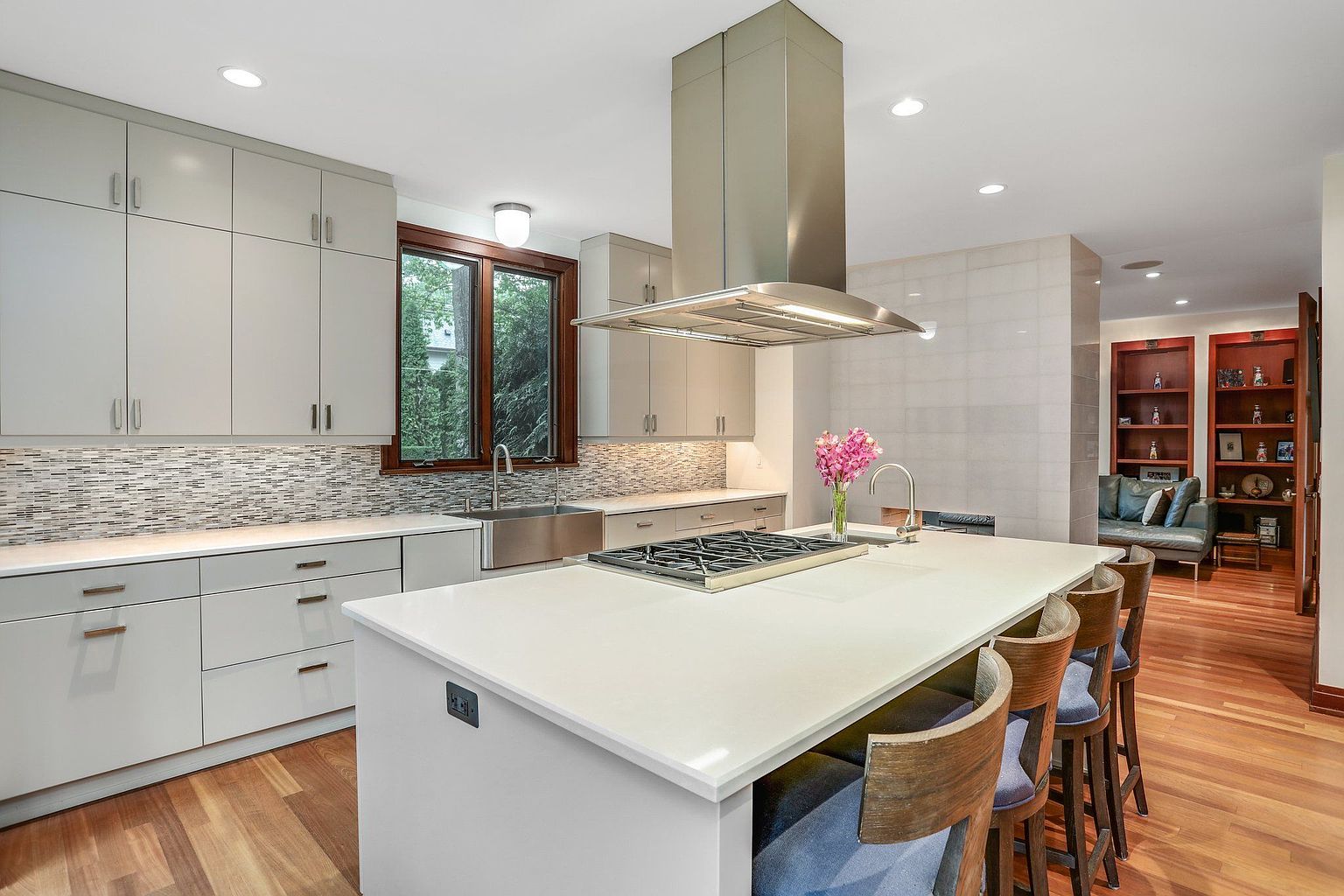 This is a well-lit, modern kitchen featuring sleek, light gray cabinetry and a large central island with a white countertop and a built-in cooktop. Stainless steel appliances and a contemporary range hood add to the modern aesthetic. The hardwood flooring and bar seating create a warm and inviting atmosphere.