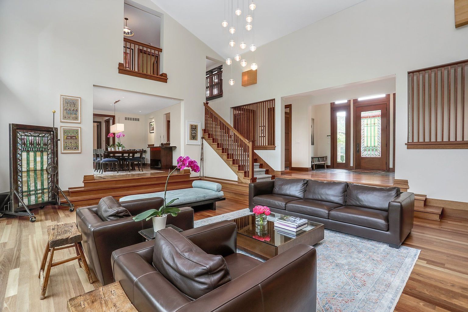 This is a spacious living room featuring hardwood floors, high ceilings, and a staircase. The room is furnished with brown leather sofas, a glass coffee table, and a decorative rug. The open layout connects to the dining area, creating a seamless flow, and the large windows and open entryway provide ample natural light.