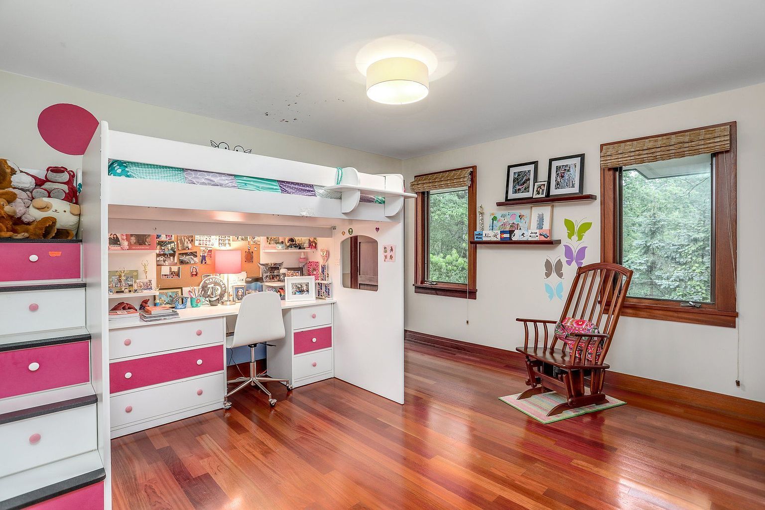 This bedroom features a unique loft bed with a built-in desk and storage, maximizing space. The room is decorated with a playful touch, including pink accents and butterfly decals. A rocking chair sits near the windows, adding a cozy element to the room.