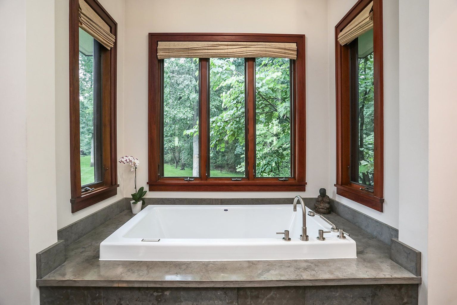 This is a well-lit primary bathroom featuring a modern white bathtub set against a gray stone platform. Three windows with wooden frames offer a view of lush greenery outside, enhancing the spa-like atmosphere. A small orchid and a Buddha statue add decorative touches to the serene space.