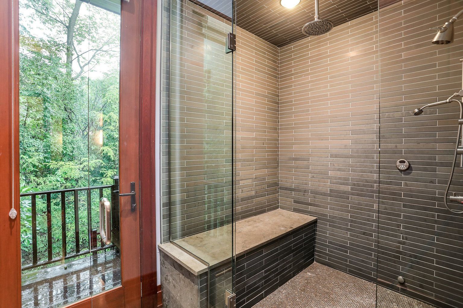 This is a modern primary bathroom featuring a walk-in shower with horizontal gray tiling and a built-in bench. The shower has a glass enclosure and a rain shower head. A door leads to a balcony with a view of lush greenery, creating a spa-like atmosphere.